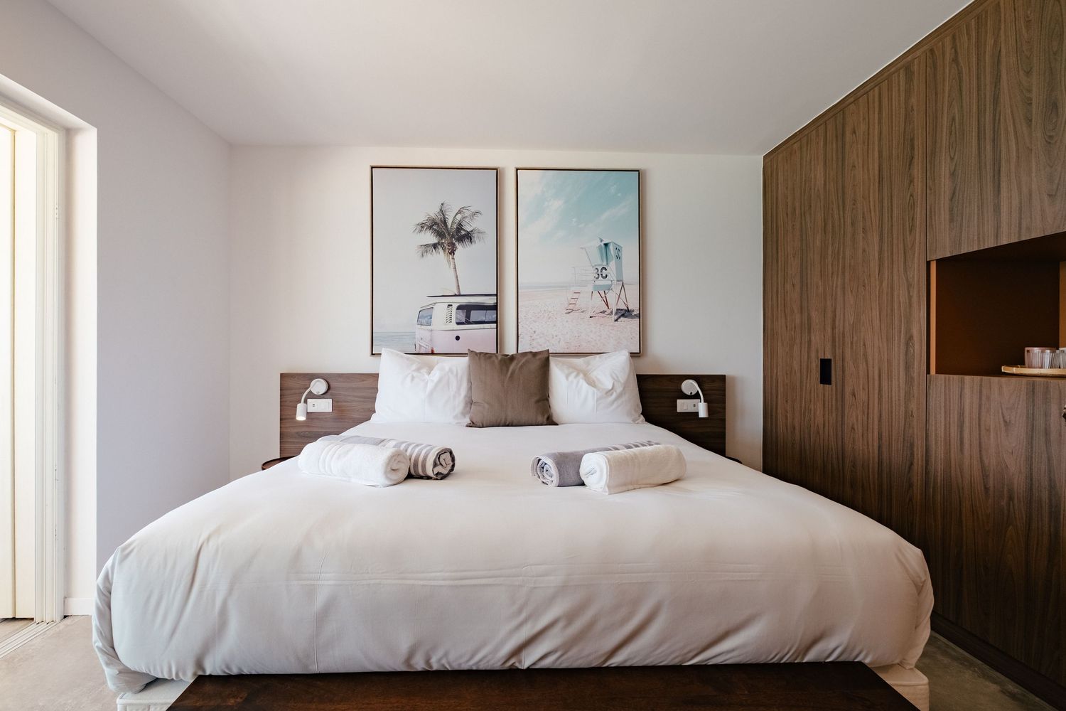 Modern hotel room interior with green velvet sofa, white bed, wooden bench and botanical artwork on neutral walls.
