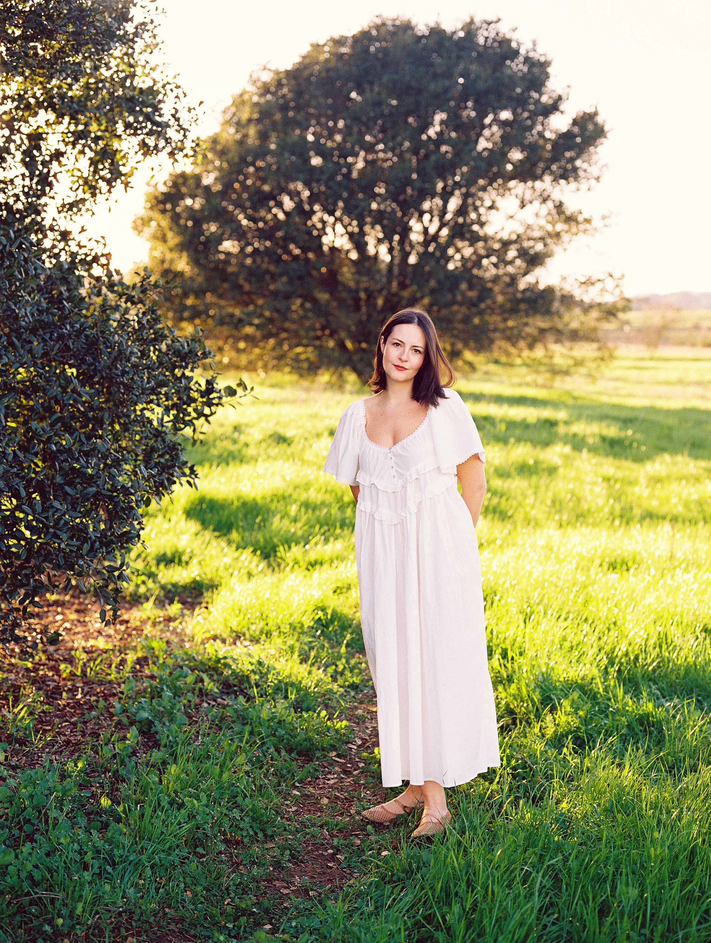 Woman in white dress standing in sunlit grassy field during golden hour.