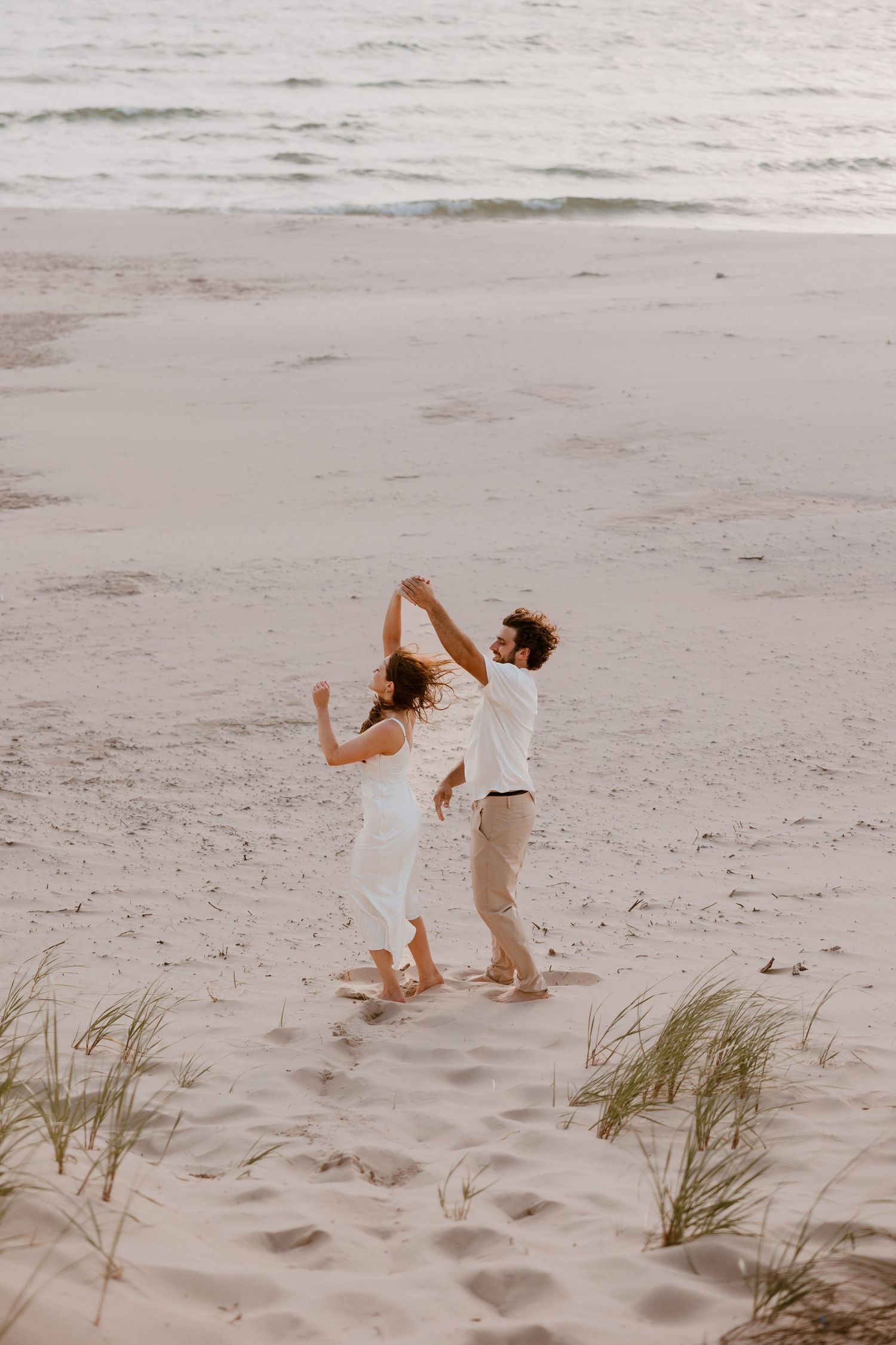 A couple dances together on a serene beach at sunset with beach grass and footprints in the sand. Michigan Wedding Photographer