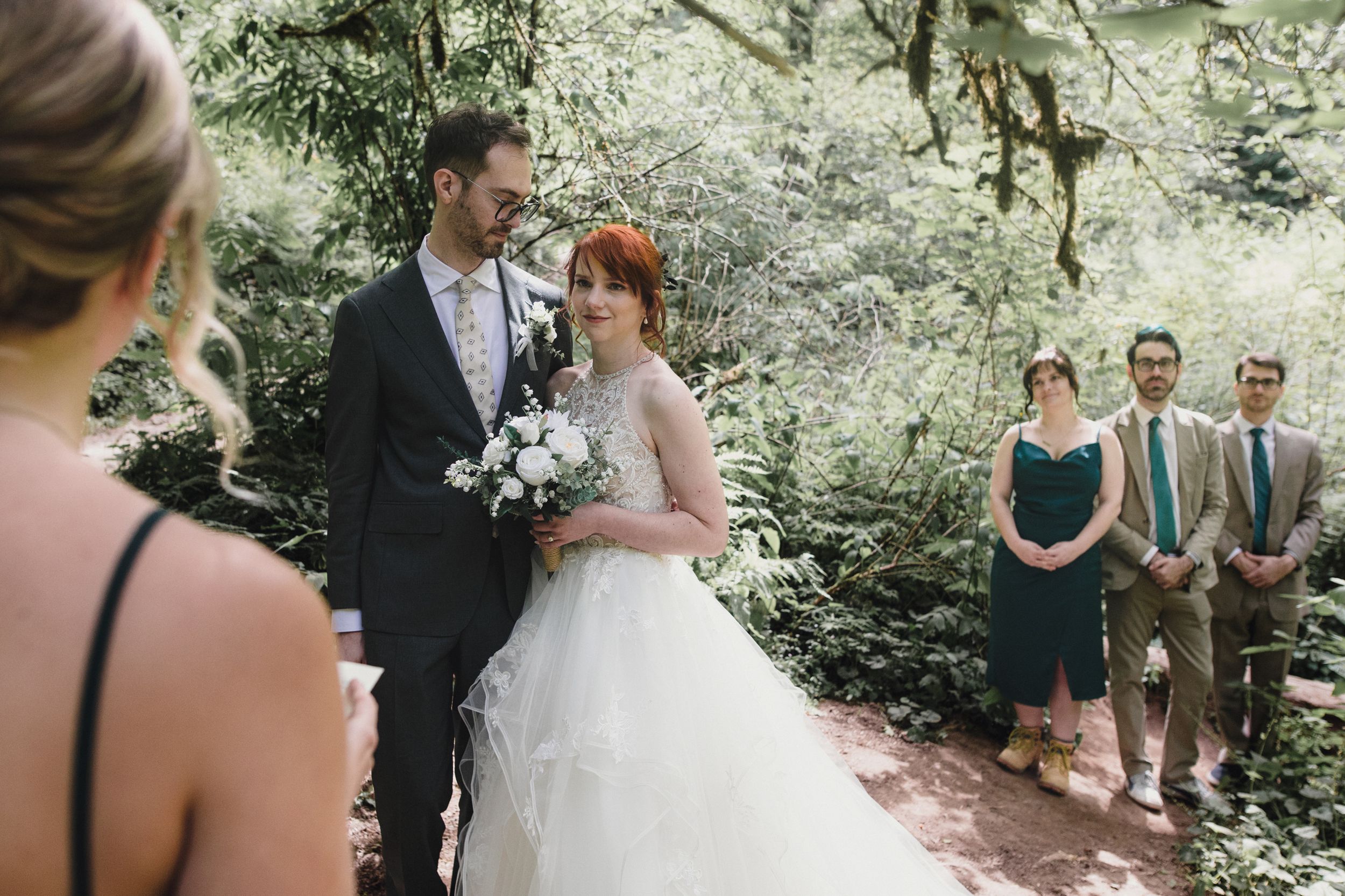 Elegant outdoor wedding ceremony in a lush forest setting with groomsmen and bridesmaids looking on in warm sunlight.