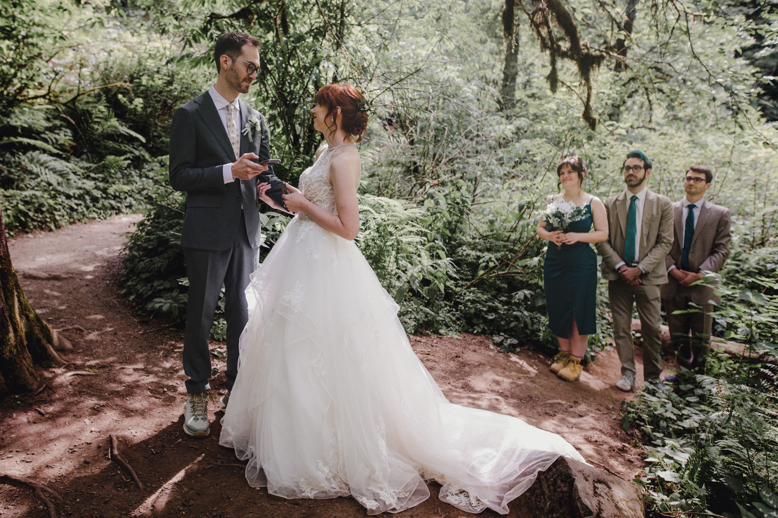 Wedding party poses together on wooded forest path surrounded by lush greenery and sunlight filtering through trees.