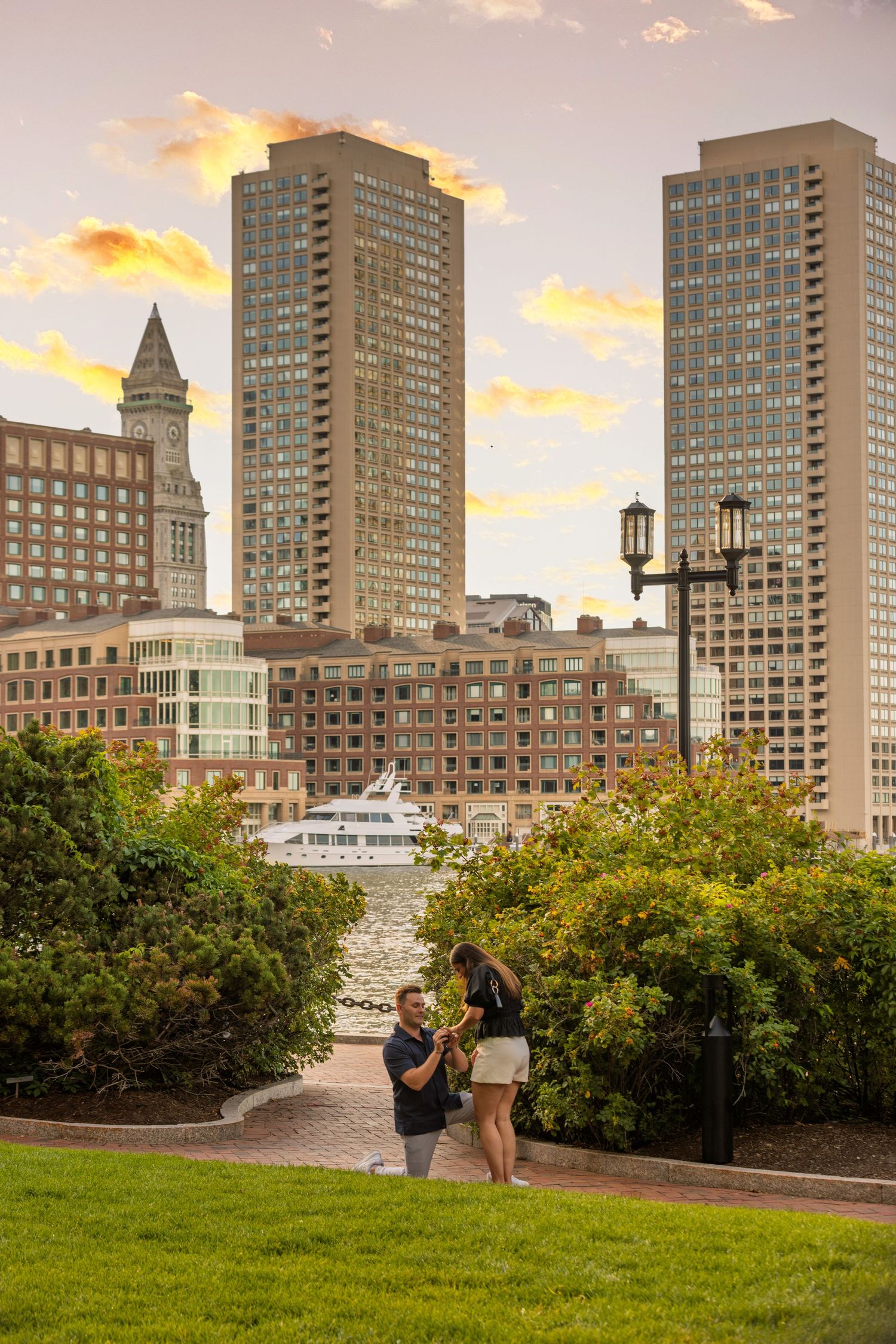 Couple enjoying sunset views of Boston skyline from a grassy park with tall skyscrapers and historic buildings in background.