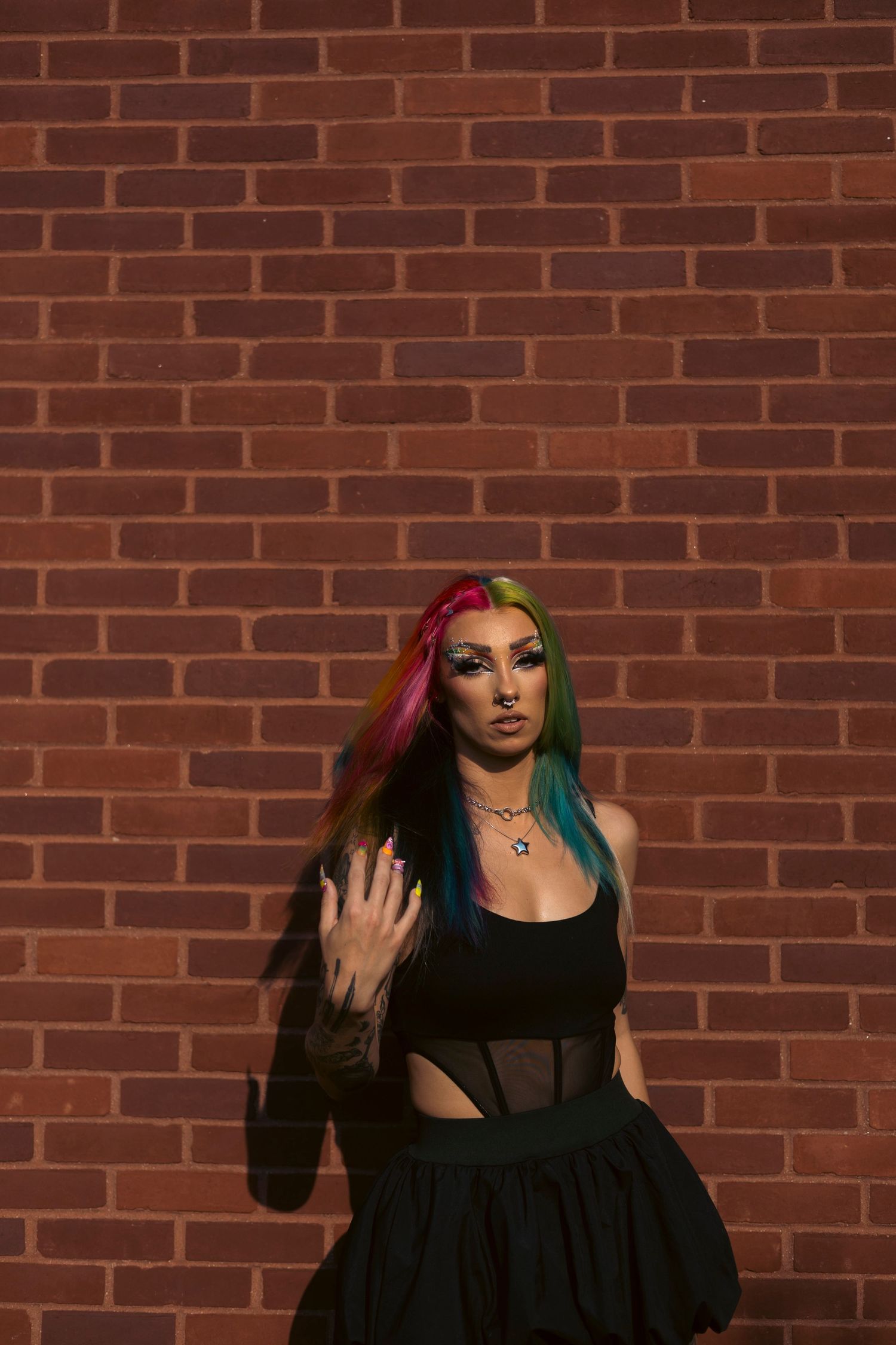 Person with vibrant multi-colored hair and black outfit poses against brick wall in artistic photo series.