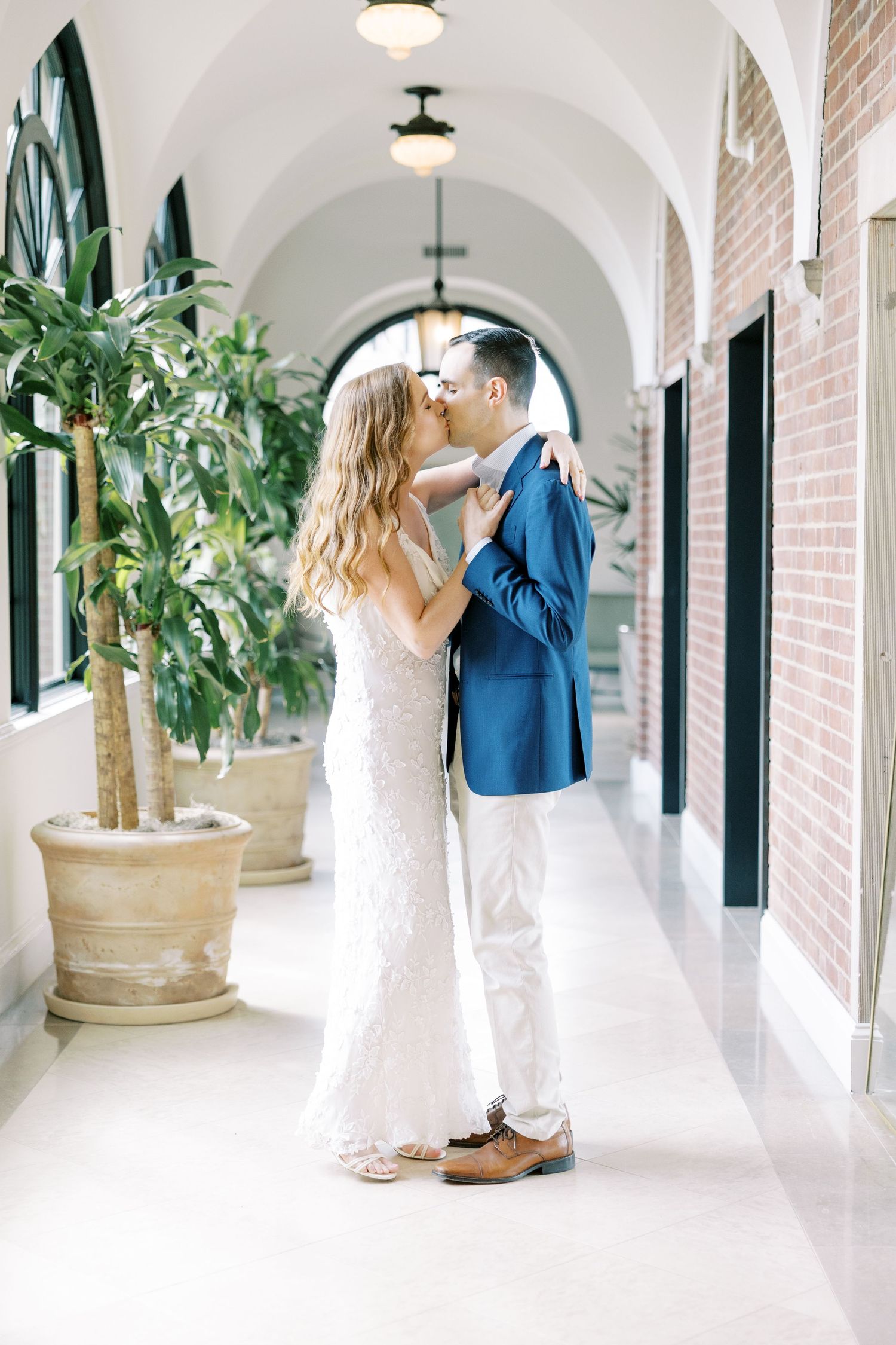 A romantic couple shares tender moments in an elegant white arched hallway with potted plants and black shutters at Pendry Natirar Peapack.
