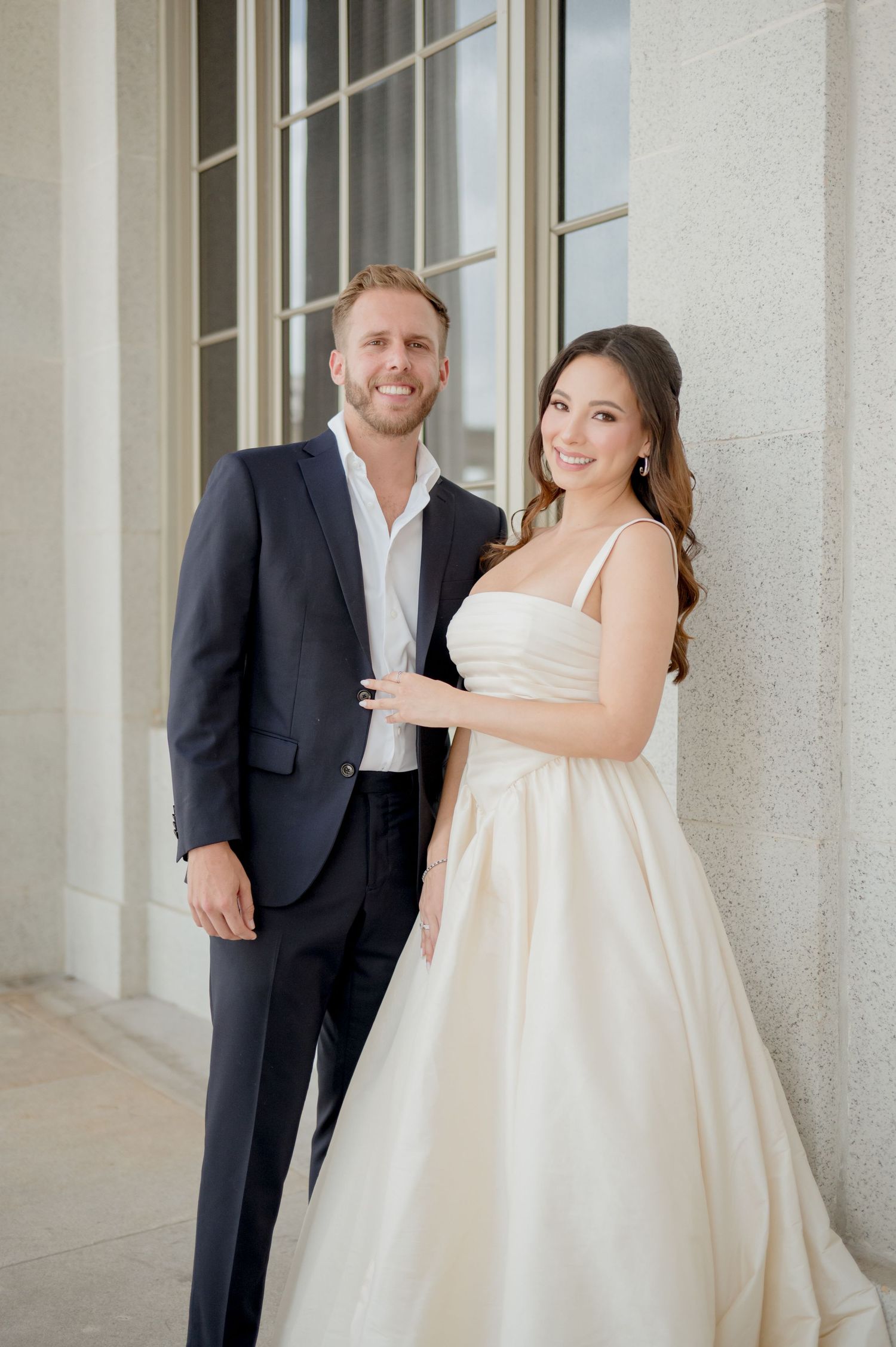 An elegant couple poses together in formal wedding attire against a white columned building exterior.