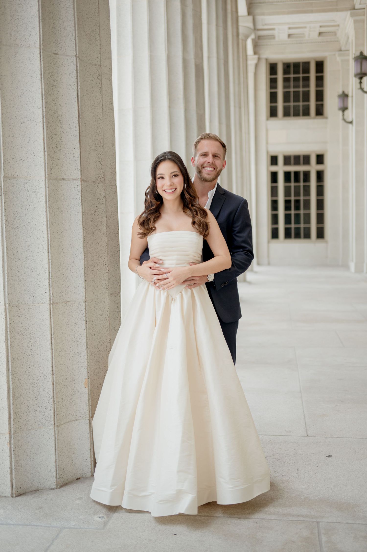 An elegant couple poses for a formal portrait between classic white columns of a grand building exterior.