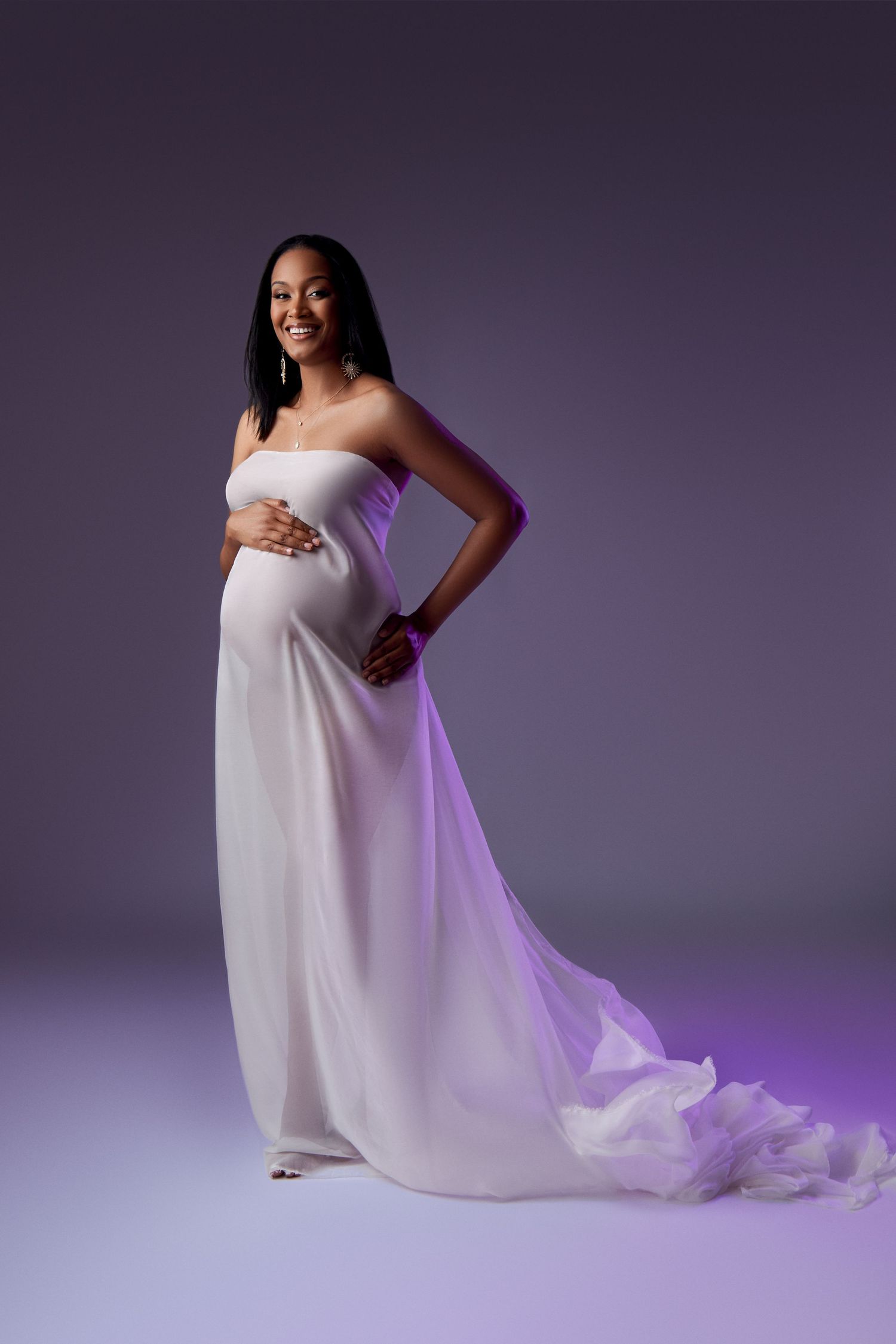 Expectant figure in flowing white gown poses gracefully against purple backdrop in elegant maternity portrait.