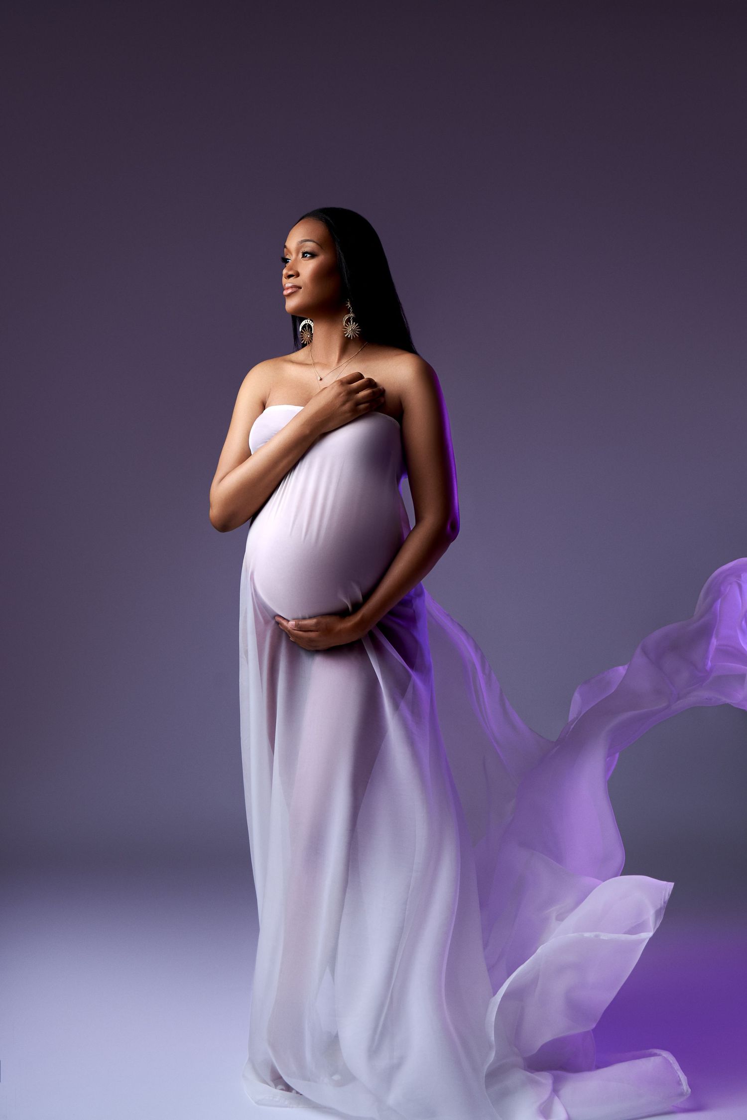Graceful maternity portrait with flowing white gown and purple fabric creating ethereal movement against dark backdrop.