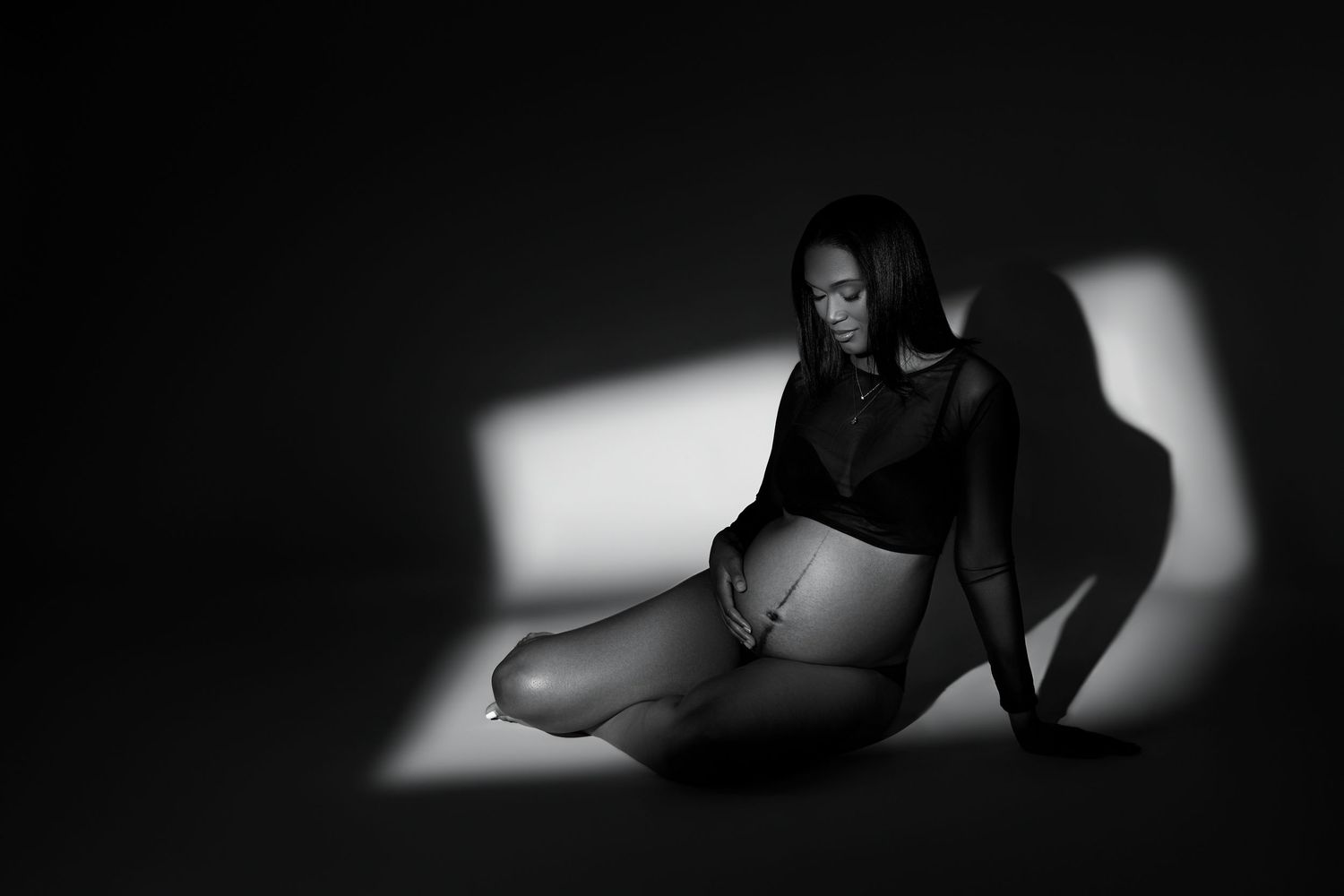 Moody black and white portrait with strong angular shadows and dramatic lighting on seated figure in dark attire.