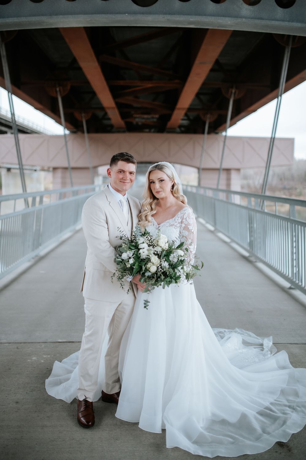 Wedding couple shares romantic moment on covered outdoor bridge walkway in white formal attire with floral bouquet.