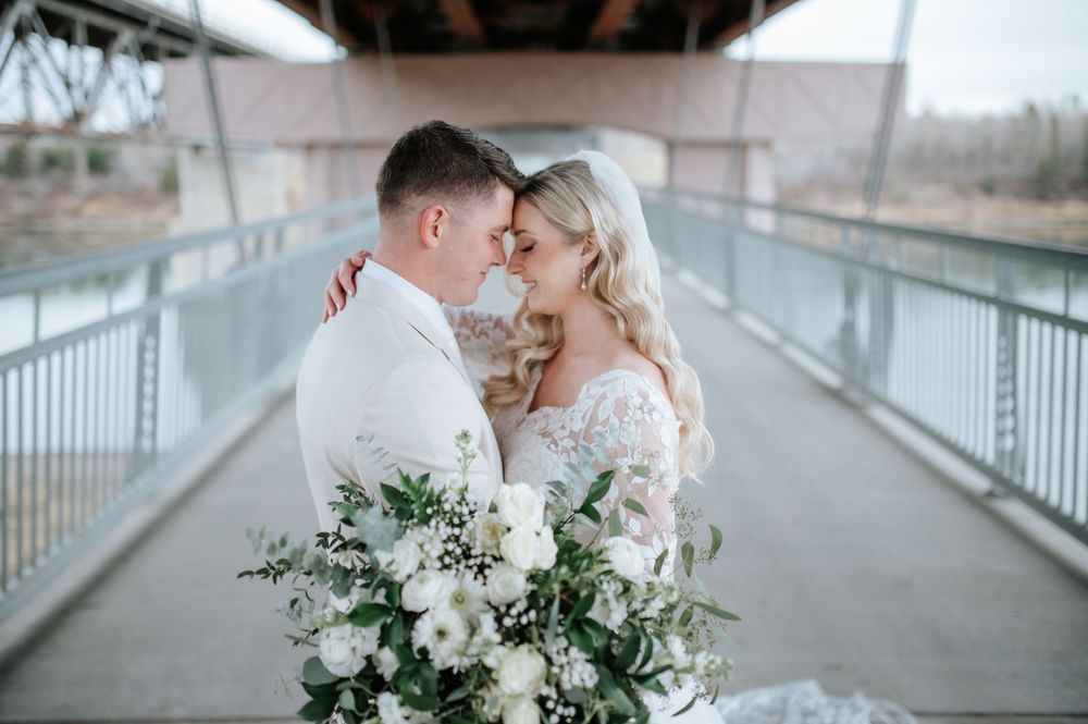 A romantic moment shared by a bride holding a white floral bouquet with greenery on a bridge walkway.