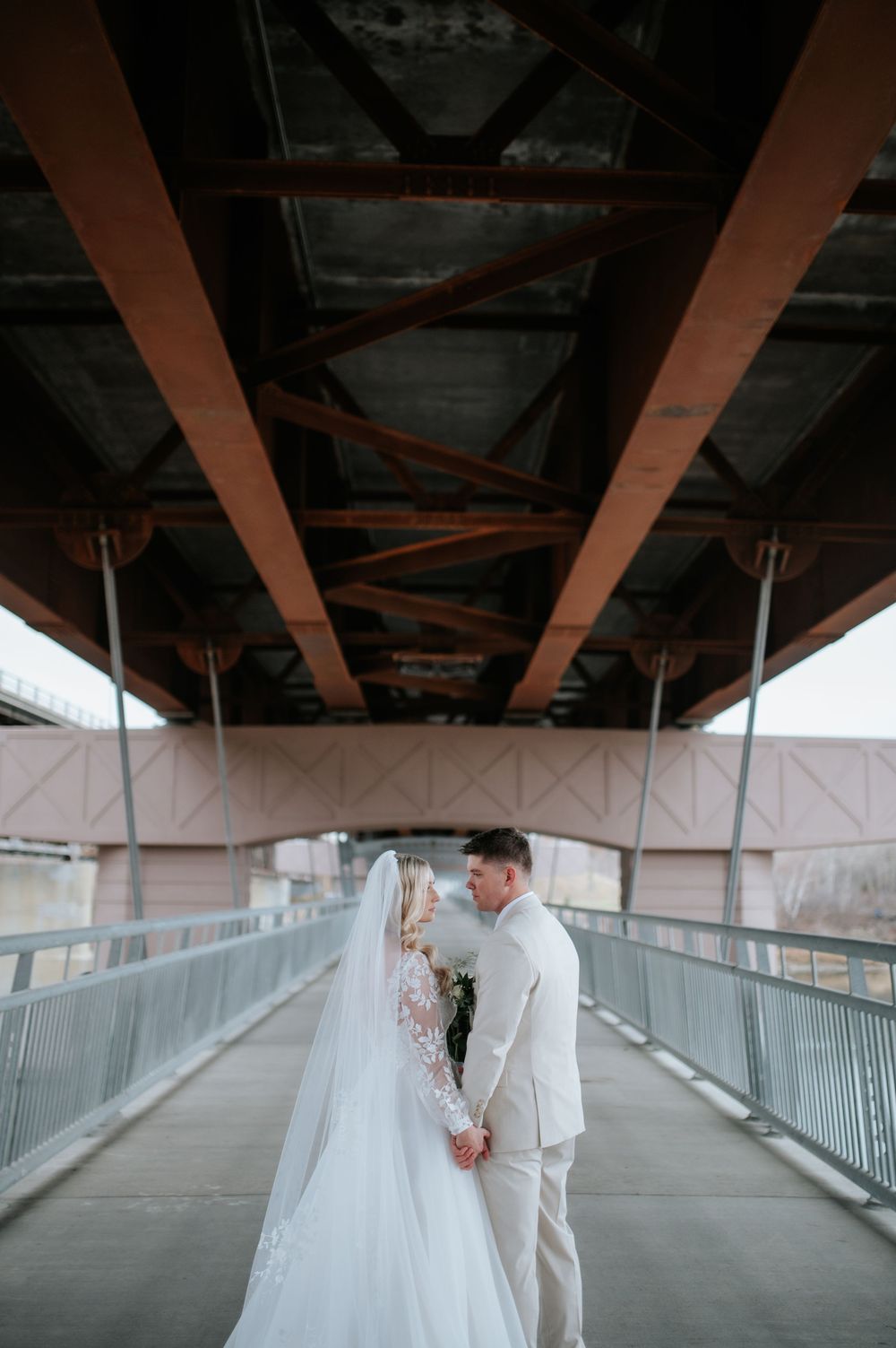 A couple in wedding attire standing on a covered bridge pathway beneath rustic steel beams.