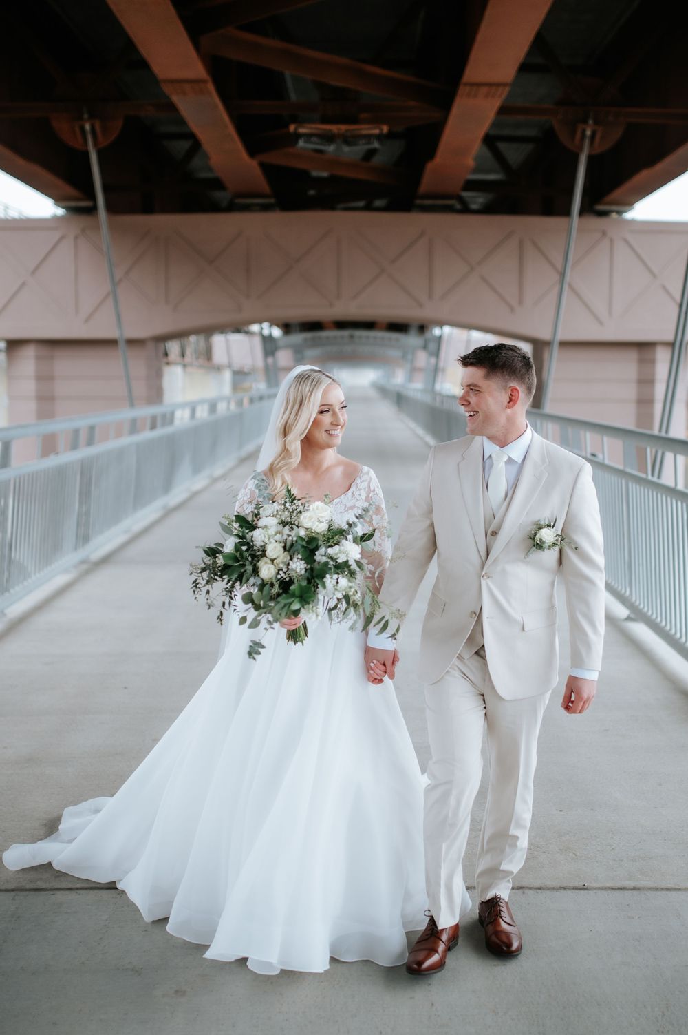 A bride in a flowing white gown and groom in a light suit walk together on a covered bridge while holding hands.