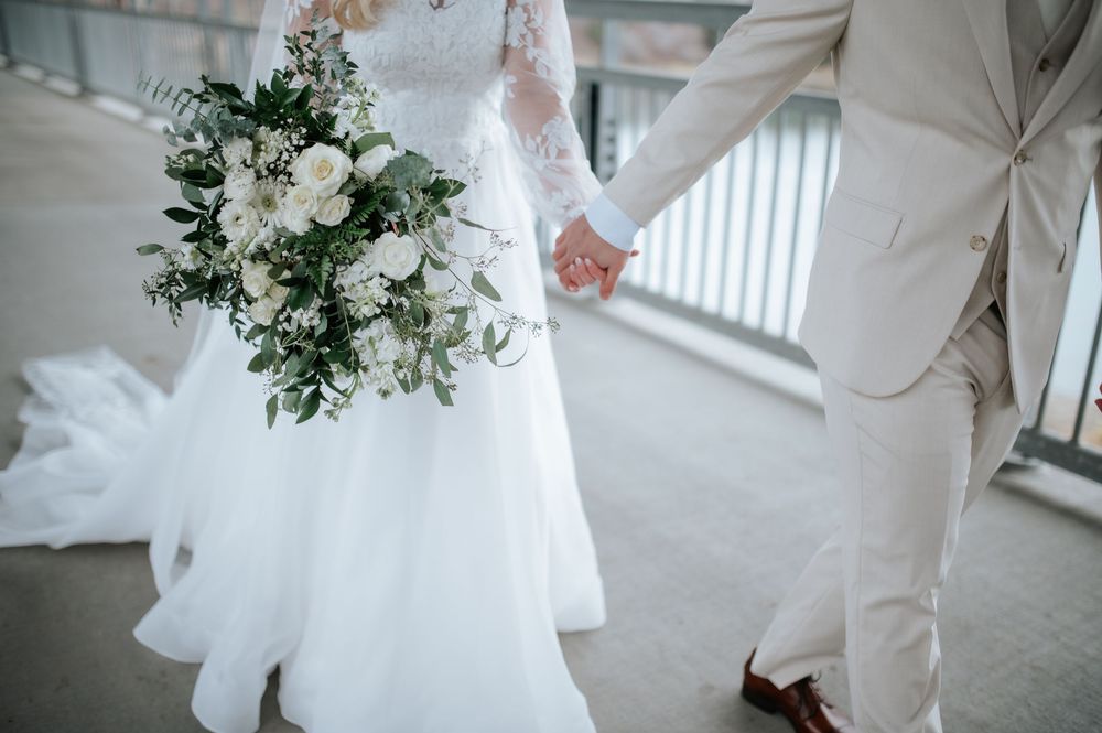 A couple stands elegantly in their white wedding attire, holding hands while walking through a bright corridor.