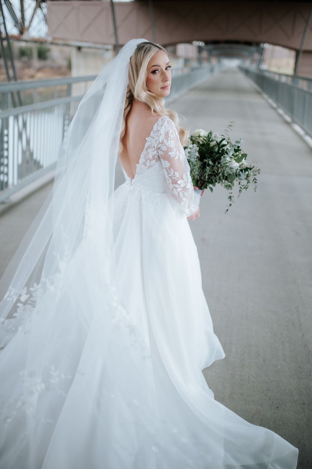 Bride in elegant lace wedding gown with long veil holds greenery bouquet while standing on outdoor walkway.