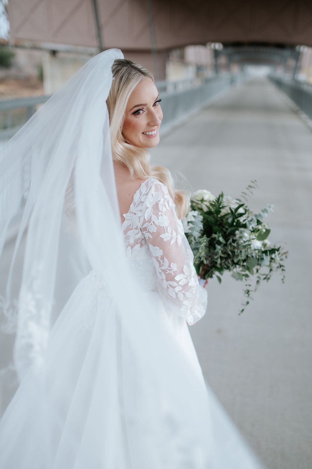 Beautiful bride in a delicate lace wedding dress with long flowing veil holding a greenery bouquet captured outdoors.