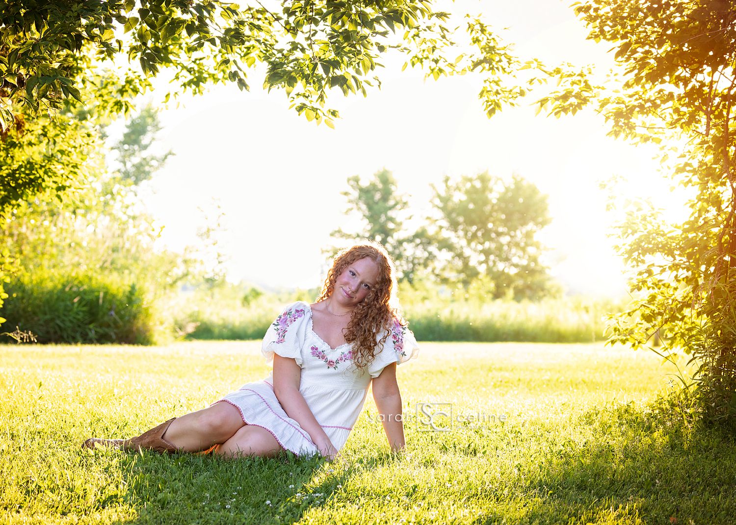 A person in a white dress sits on sunlit grass during golden hour as warm summer light filters through trees.