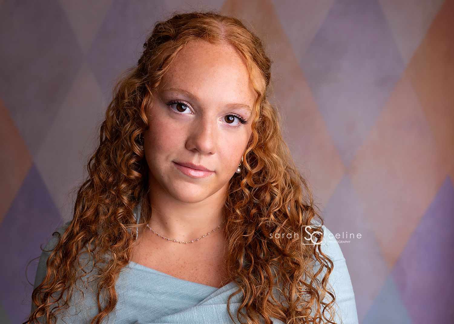 Portrait of curly haired young person in teal top against textured diamond pattern backdrop.