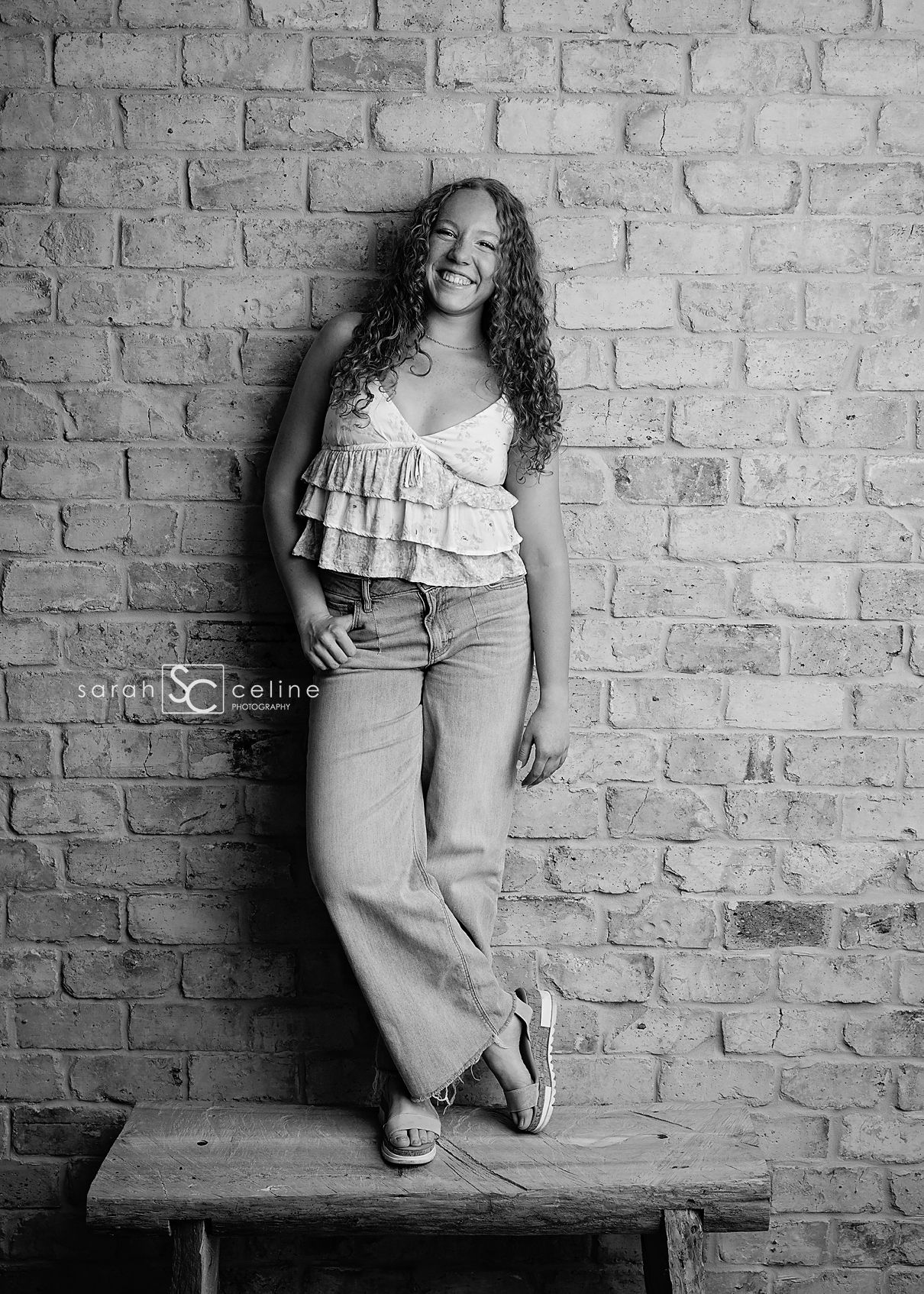 Black and white portrait of someone with curly hair wearing a ruffled top and wide-leg pants posing on bench against brick wall.