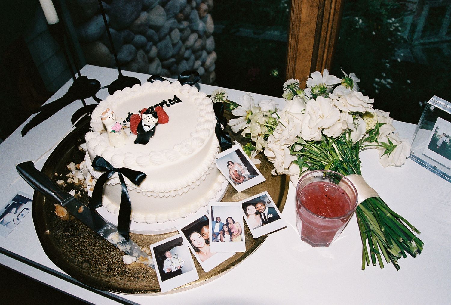 A white tiered wedding cake with black ribbon accent sits on a table alongside floral arrangements and photographs.