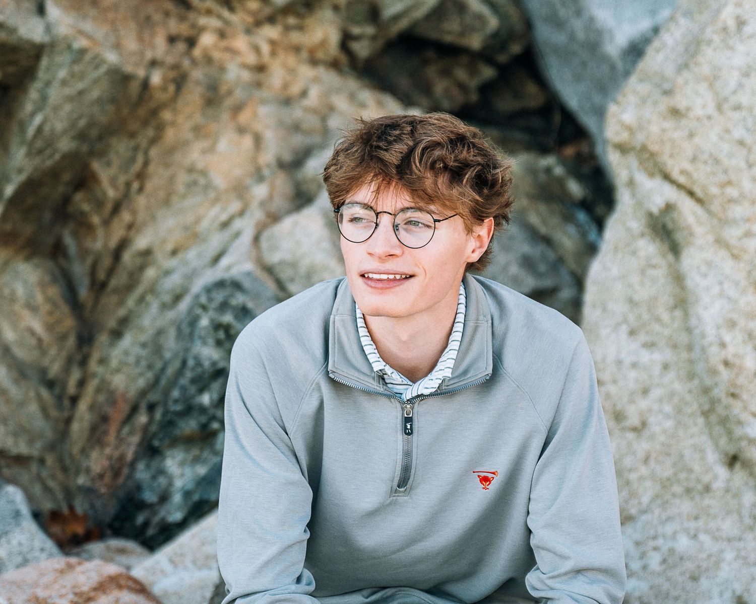 Casual outdoor portrait with glasses and grey half-zip sweater against rocky background.
