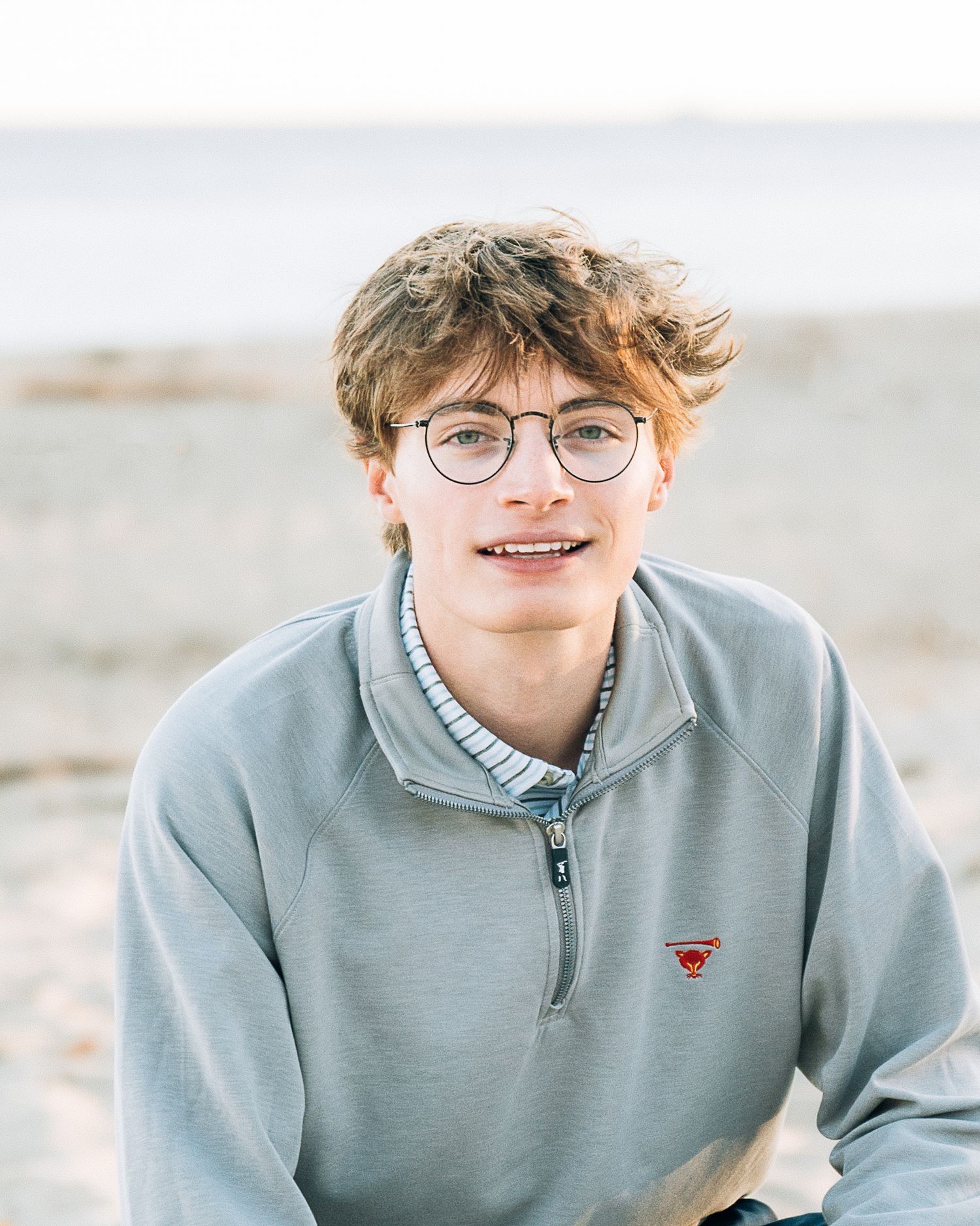 Close-up portrait wearing glasses and grey zip-up sweater against coastal scenery.