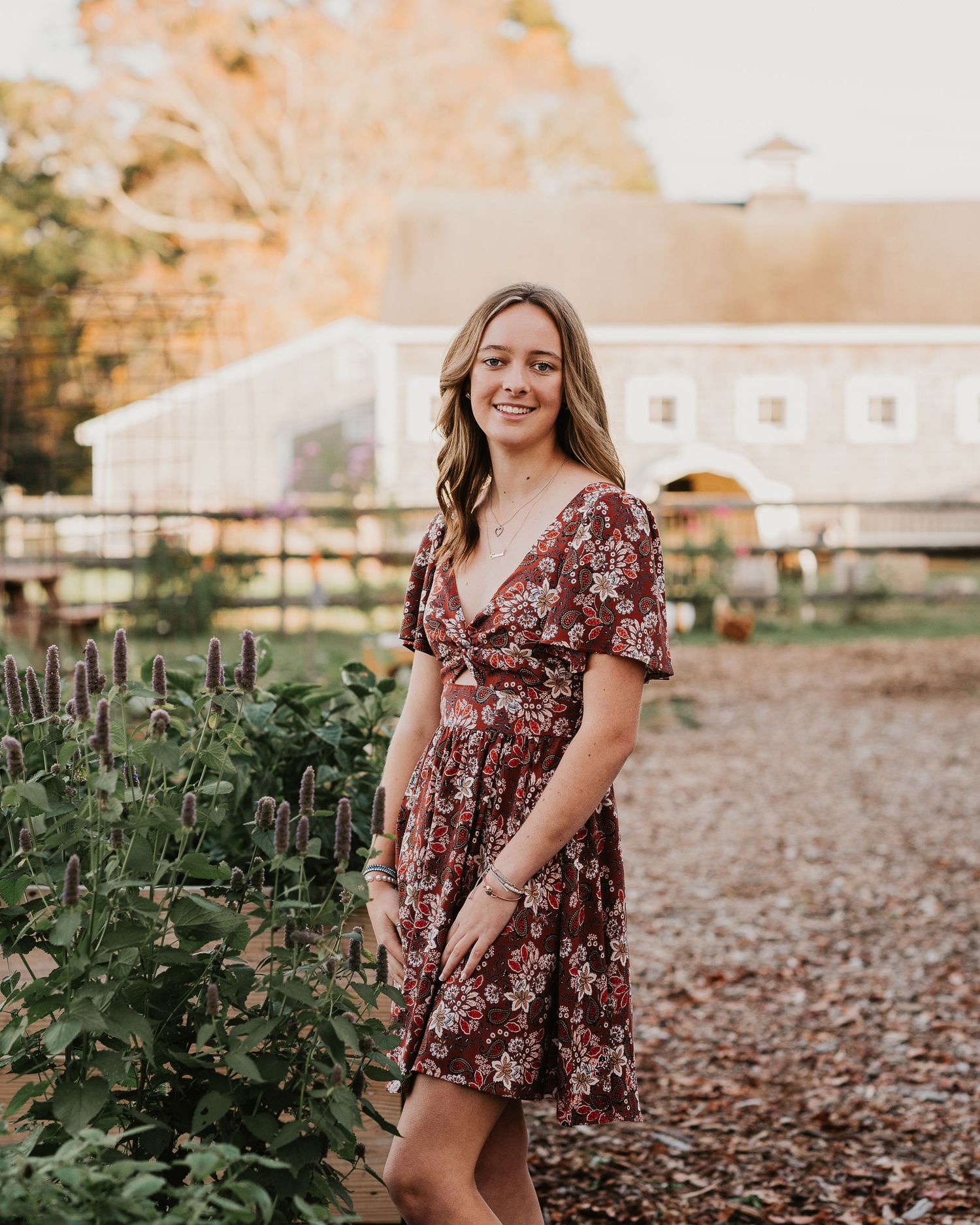 Portrait in floral burgundy dress standing by garden with barn in background.