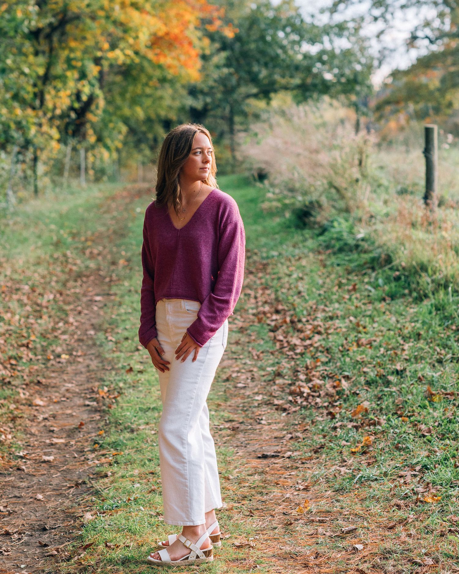 Purple sweater and white pants outfit on autumn trail with colorful foliage.