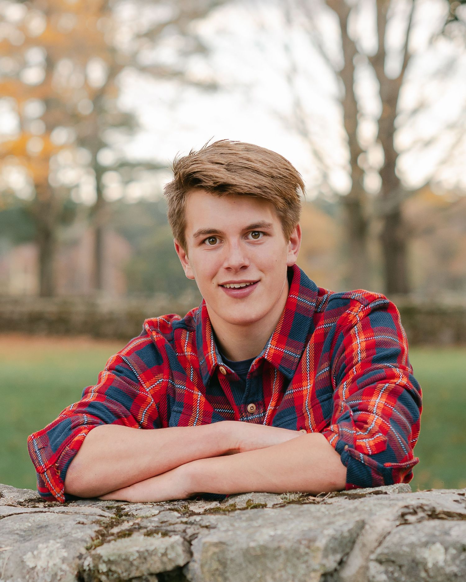 Young person in red plaid flannel shirt leaning on stone wall in autumn setting.