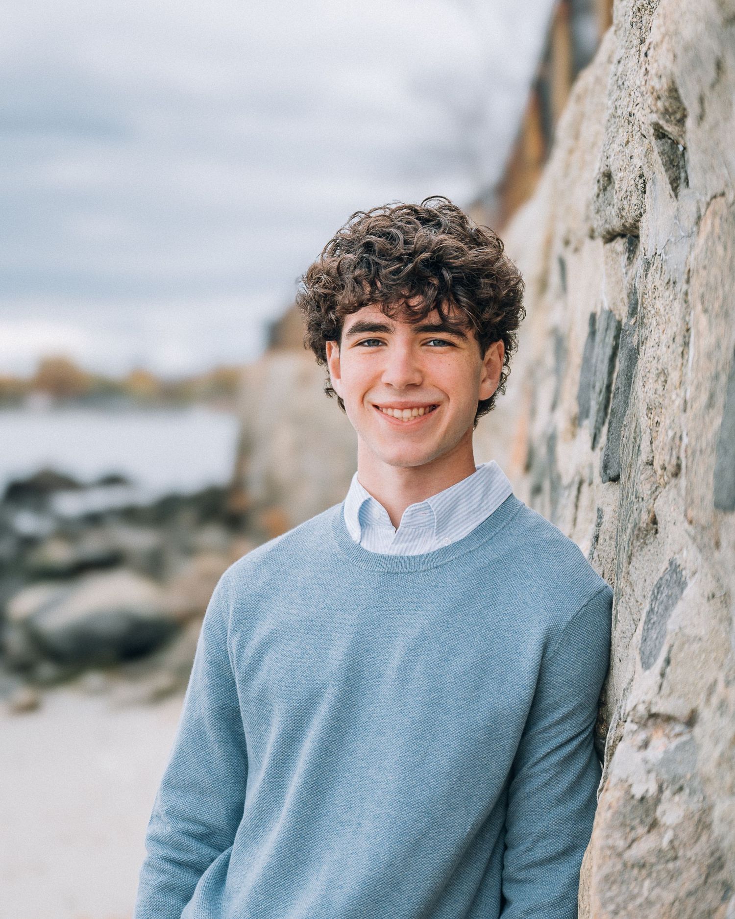 Casual portrait by a stone wall at a coastal location showing blue sweater and curly hair.