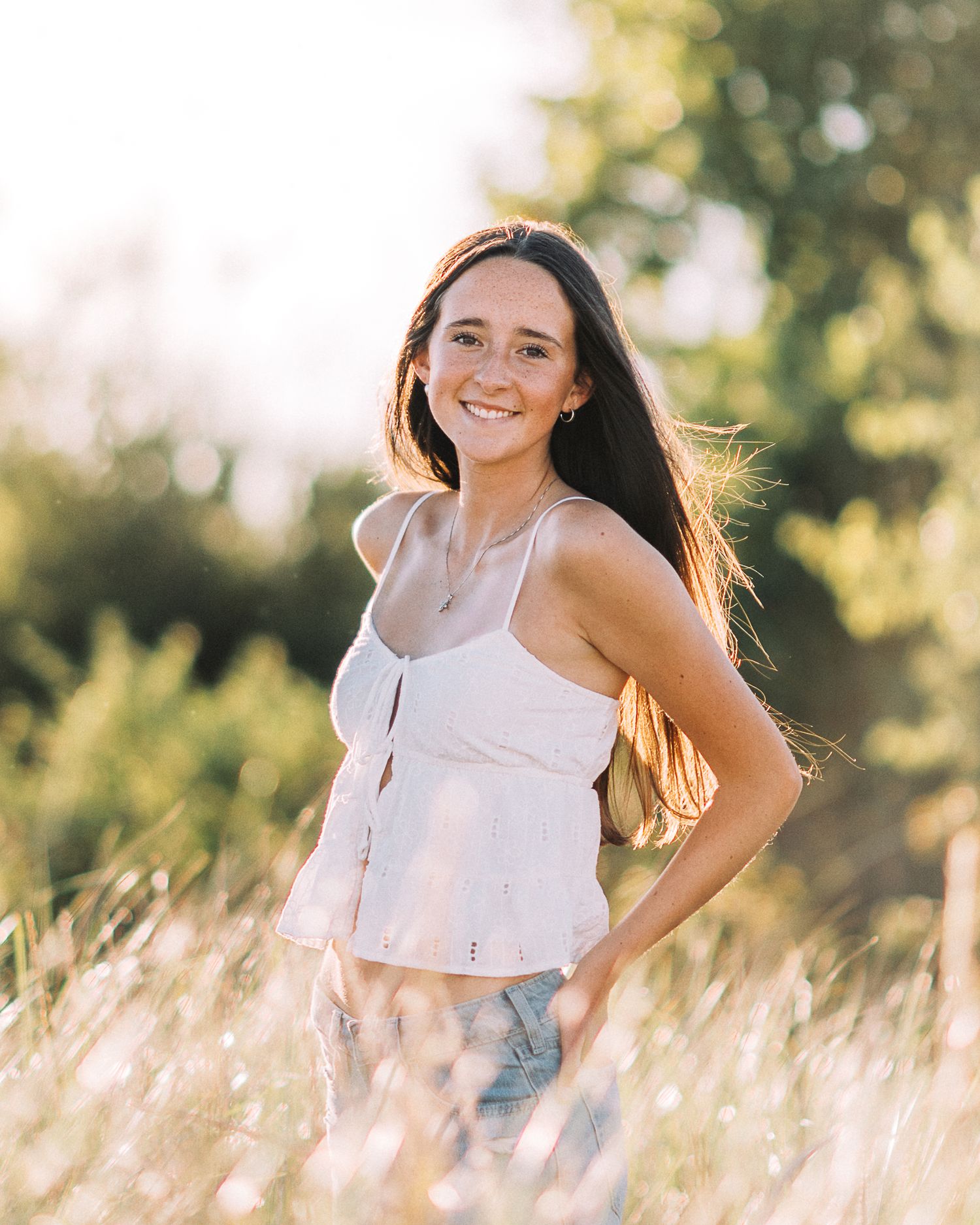 Young woman with long dark hair smiles in golden field, wearing white crop top and jeans in warm sunlight.