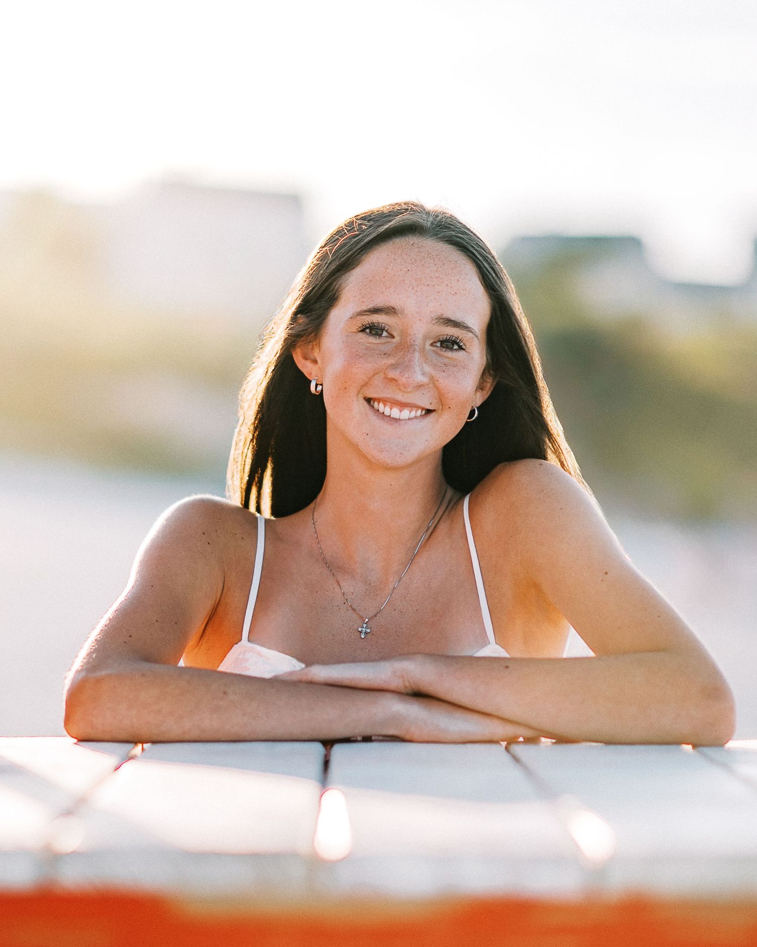 Smiling young woman with freckles leaning on a wooden surface outdoors in warm sunlight wearing a white top.