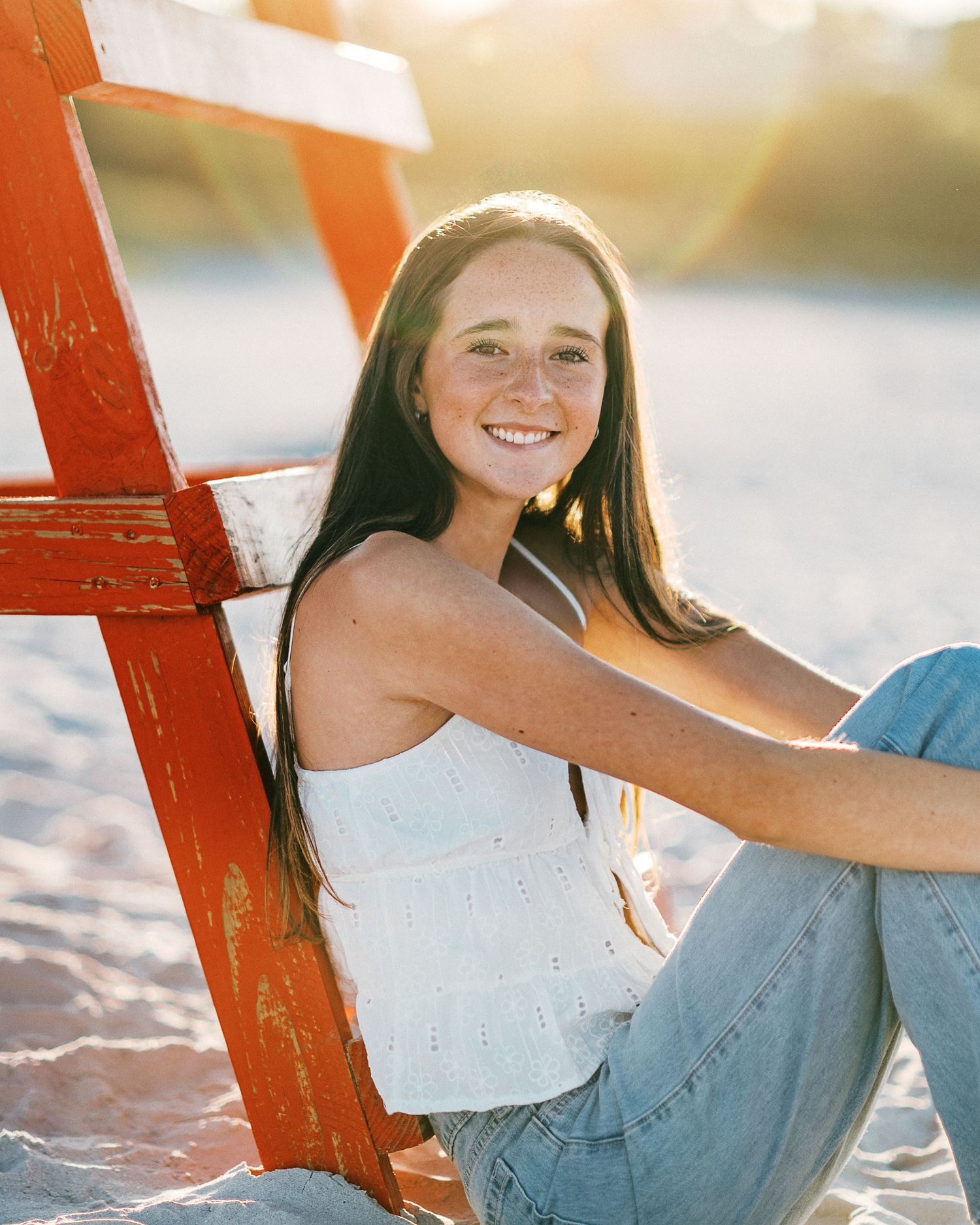 Young woman with freckles smiling on sandy beach, leaning against red lifeguard stand in white top and jeans.