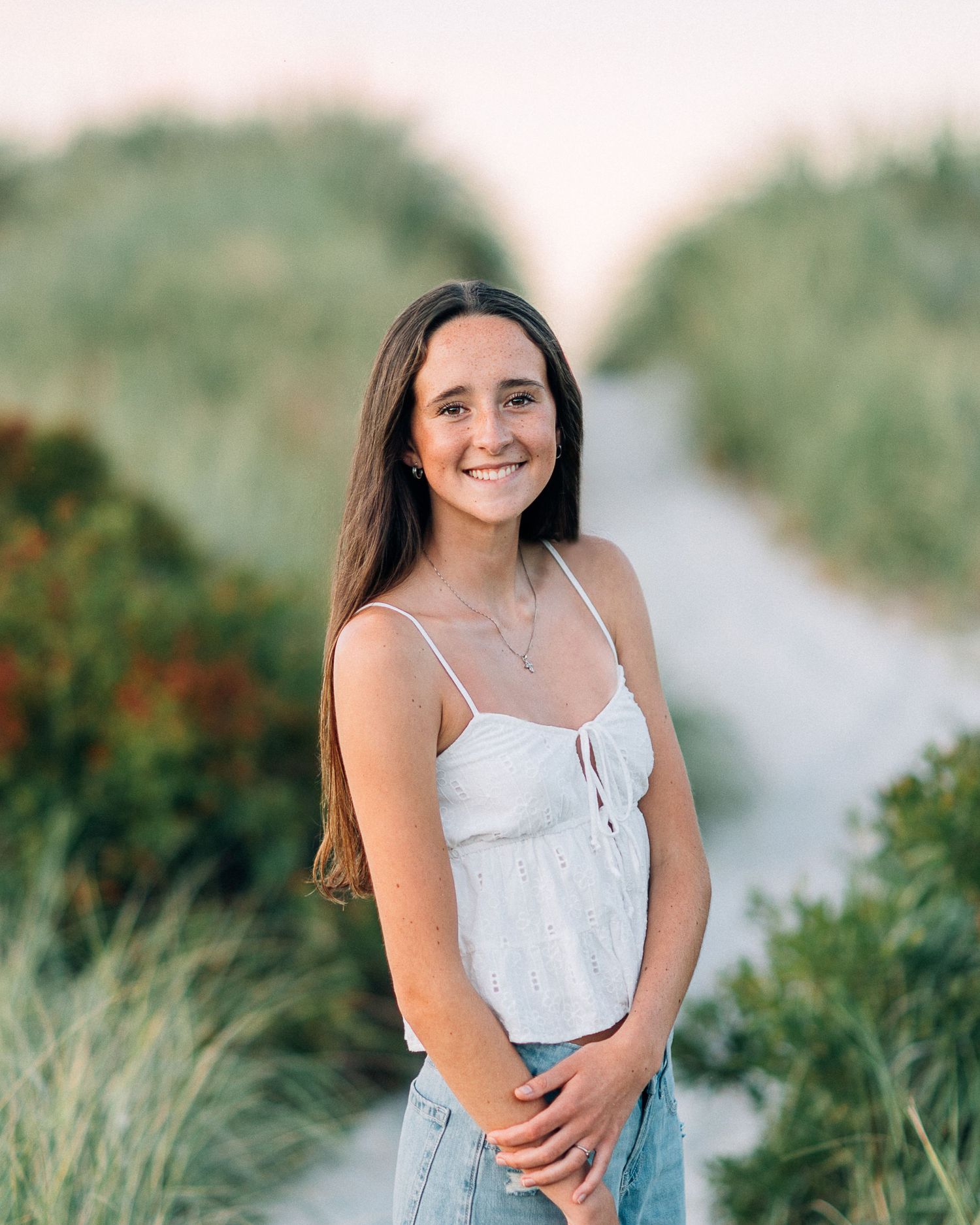 Young woman smiling on a sandy beach path, wearing a white camisole top and jeans, surrounded by green coastal plants.
