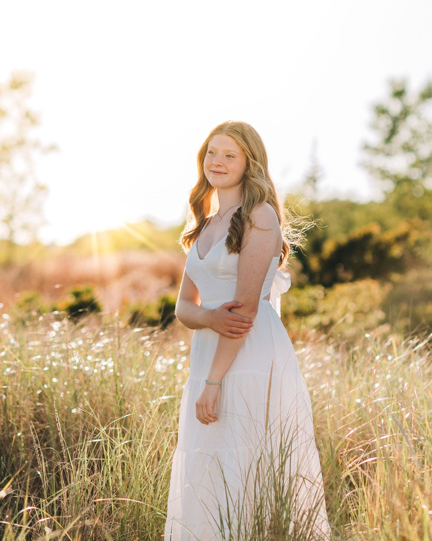 Young woman in white dress standing in golden sunlit field, smiling gracefully among wildflowers.