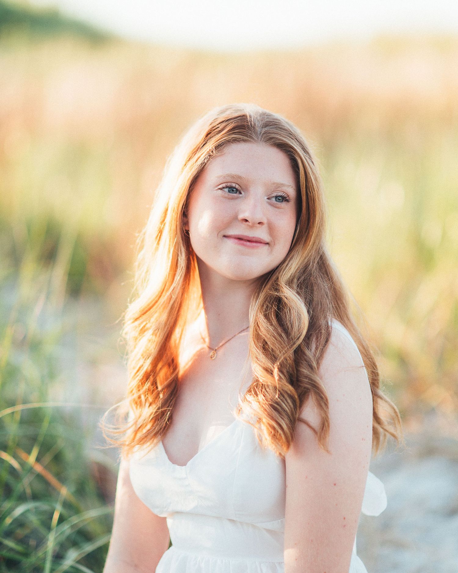 Young girl with long red hair smiling outdoors in white dress at golden hour beach setting.