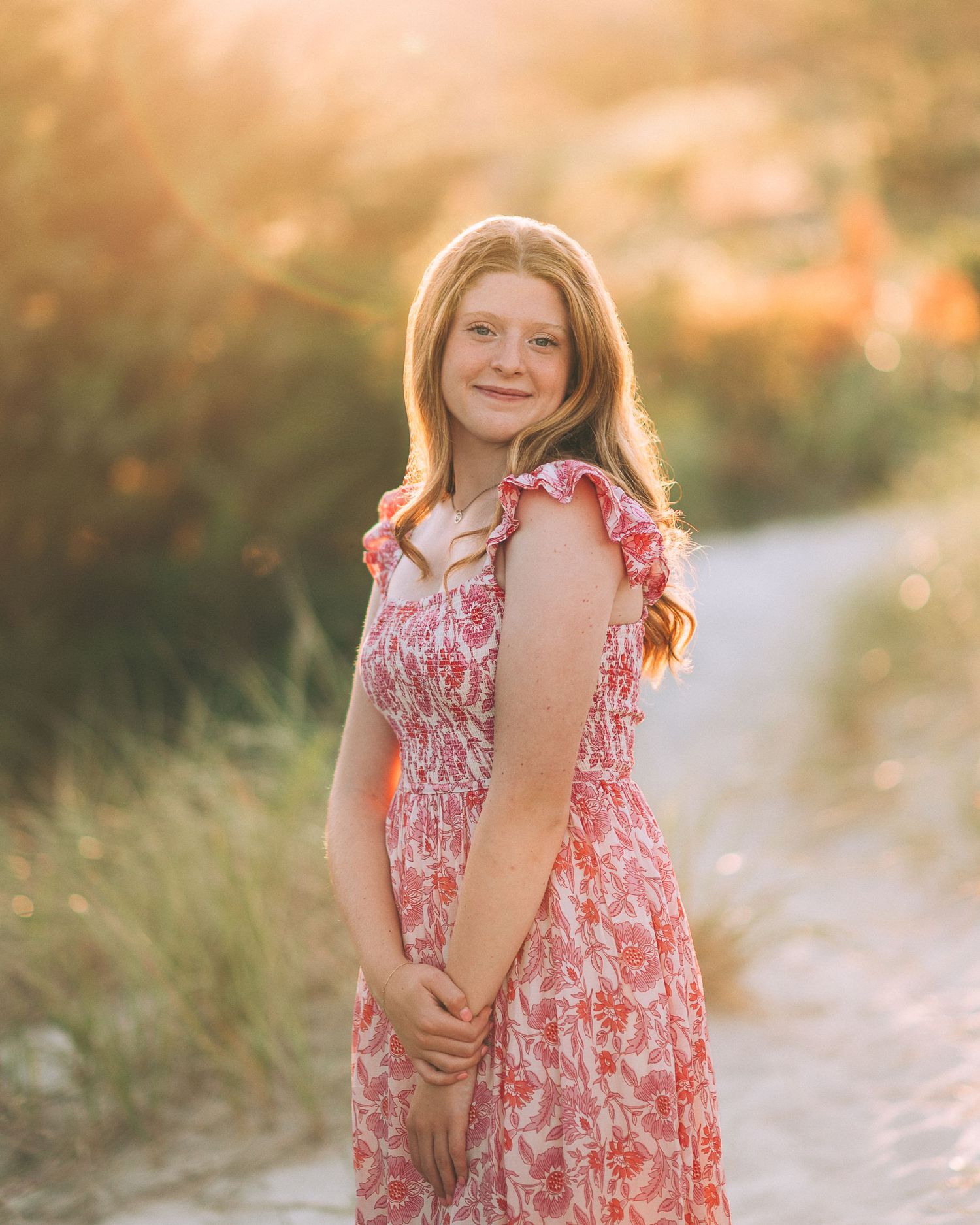 Young girl in pink floral dress smiling outdoors on a sunlit path surrounded by green grass.