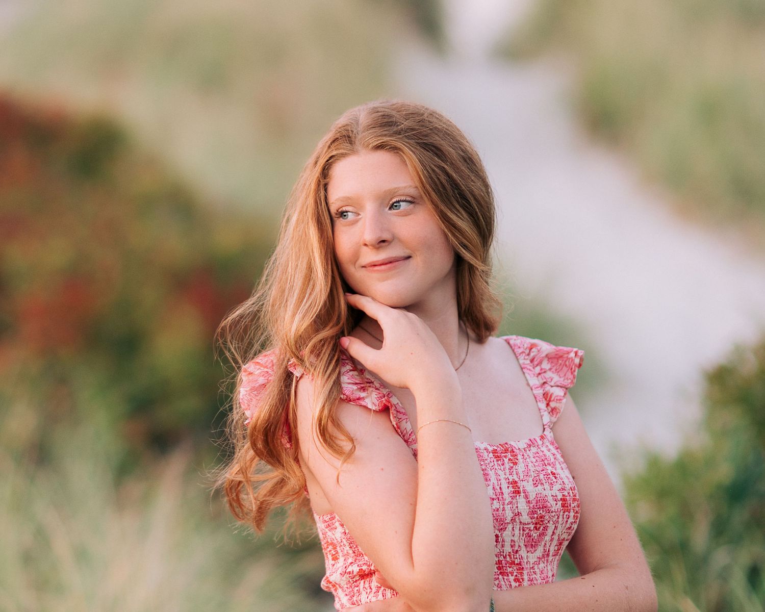 Young woman with long red hair in a pink floral dress poses outdoors in a sunny green field.