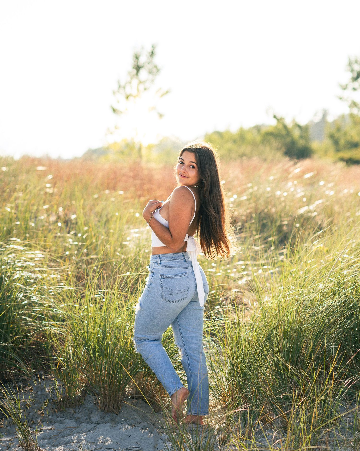 Fashion portrait in white top and jeans standing among tall grass at golden hour.
