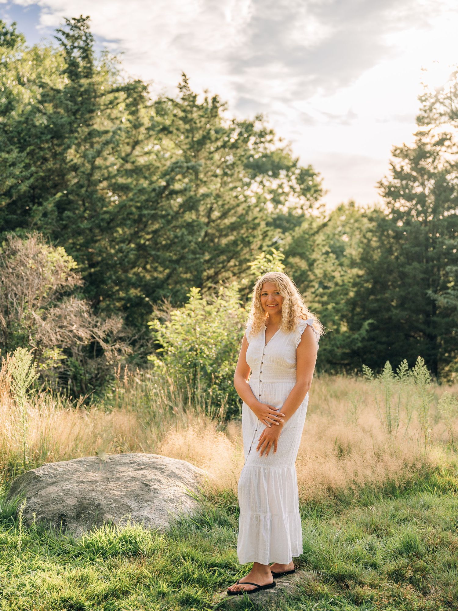 White maxi dress captured in golden sunlit meadow with trees in background.