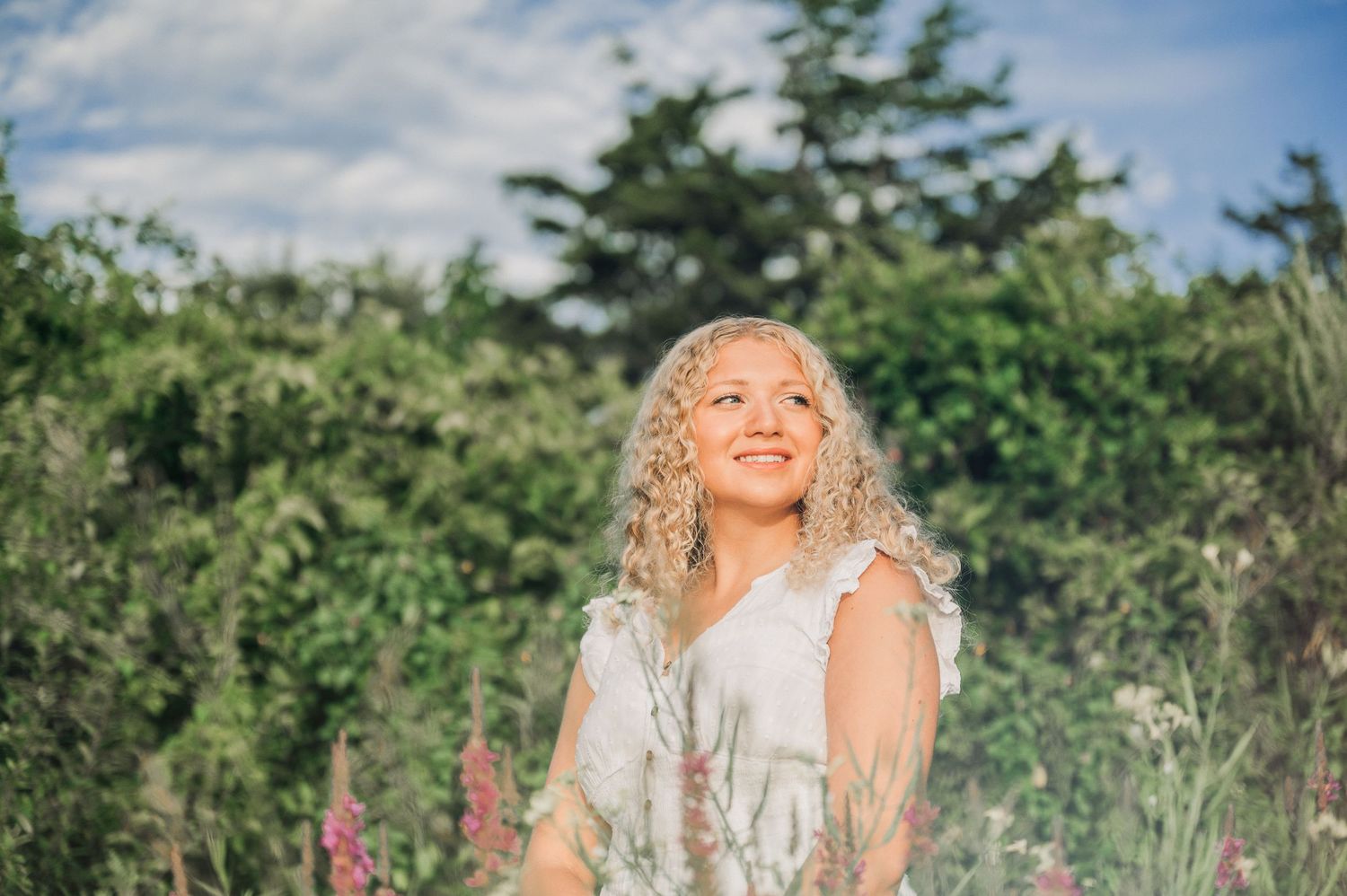 Summer portrait in white dress amid lush garden greenery under blue sky.
