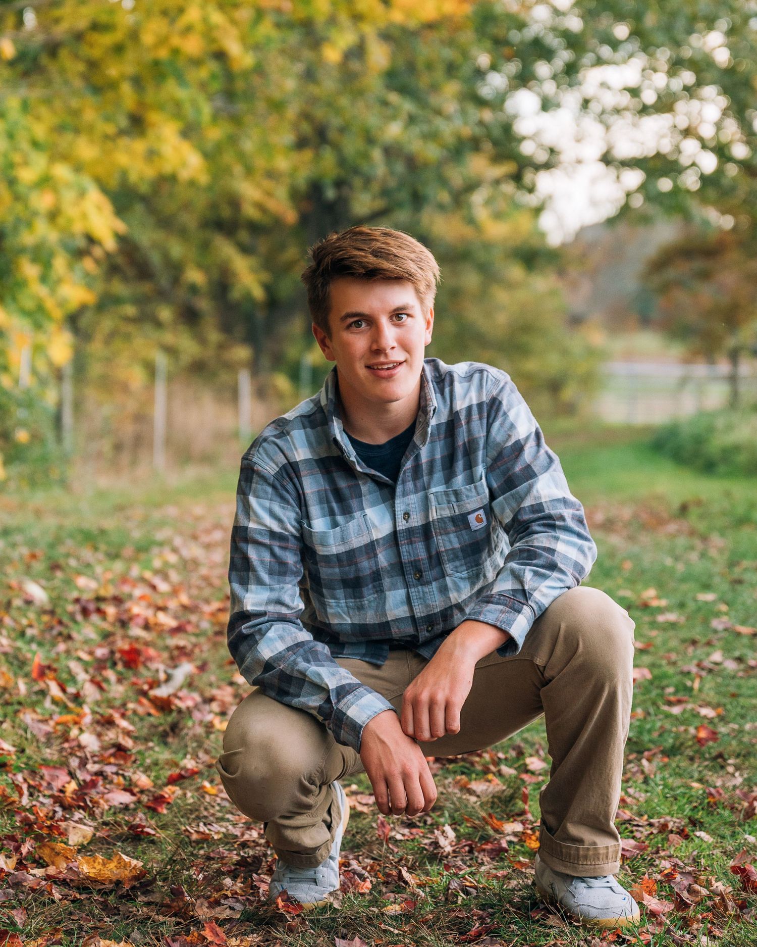 Casual portrait in plaid flannel shirt crouching among autumn leaves with trees in background.