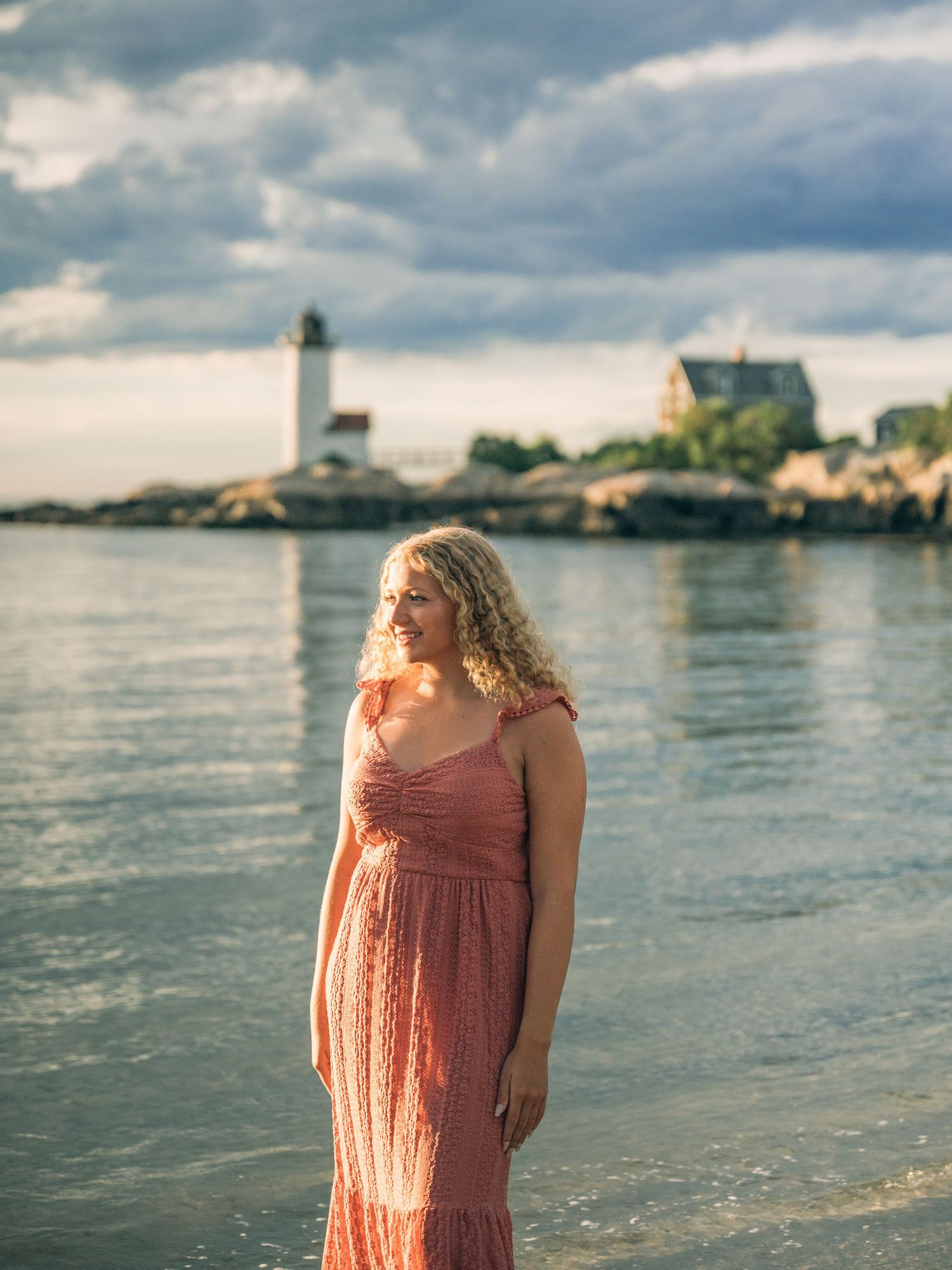Person in a coral dress poses at sunset by the ocean with coastal buildings in the background.