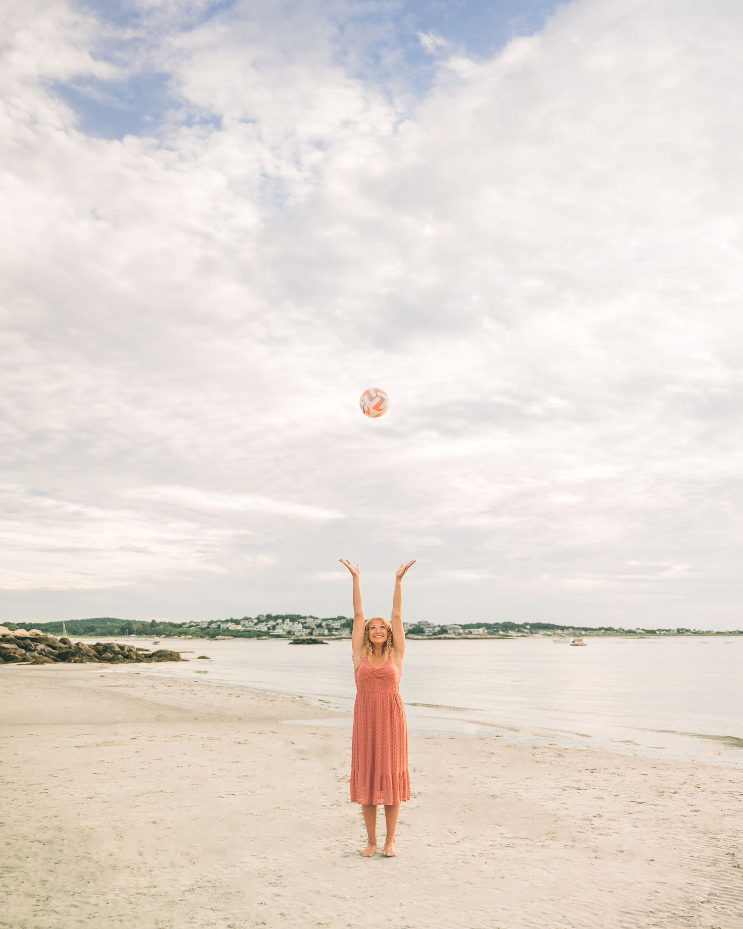 Person in a peach dress stands joyfully on an empty beach with arms raised.