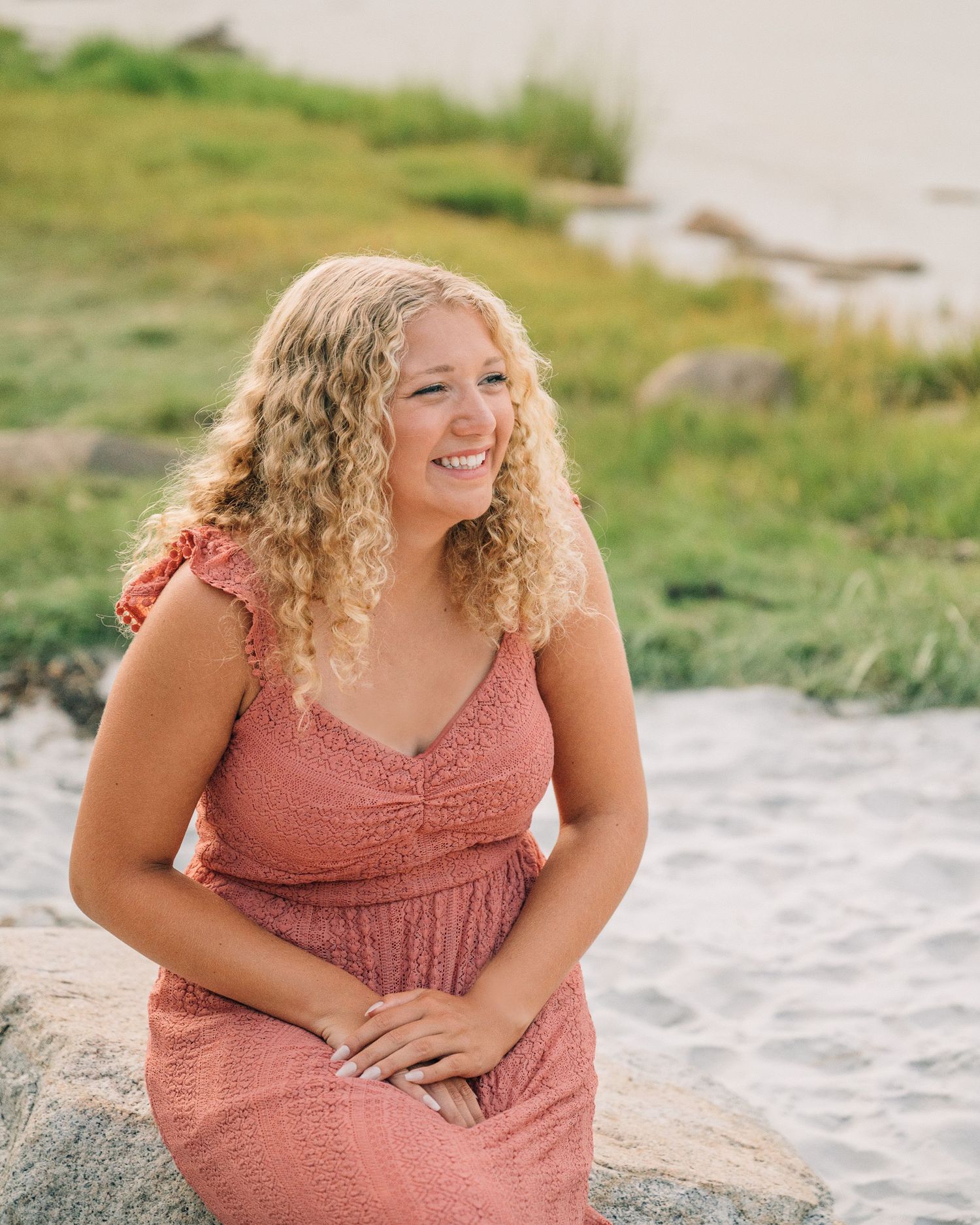 Person in a dusty rose dress sits casually on coastal rocks near the ocean.