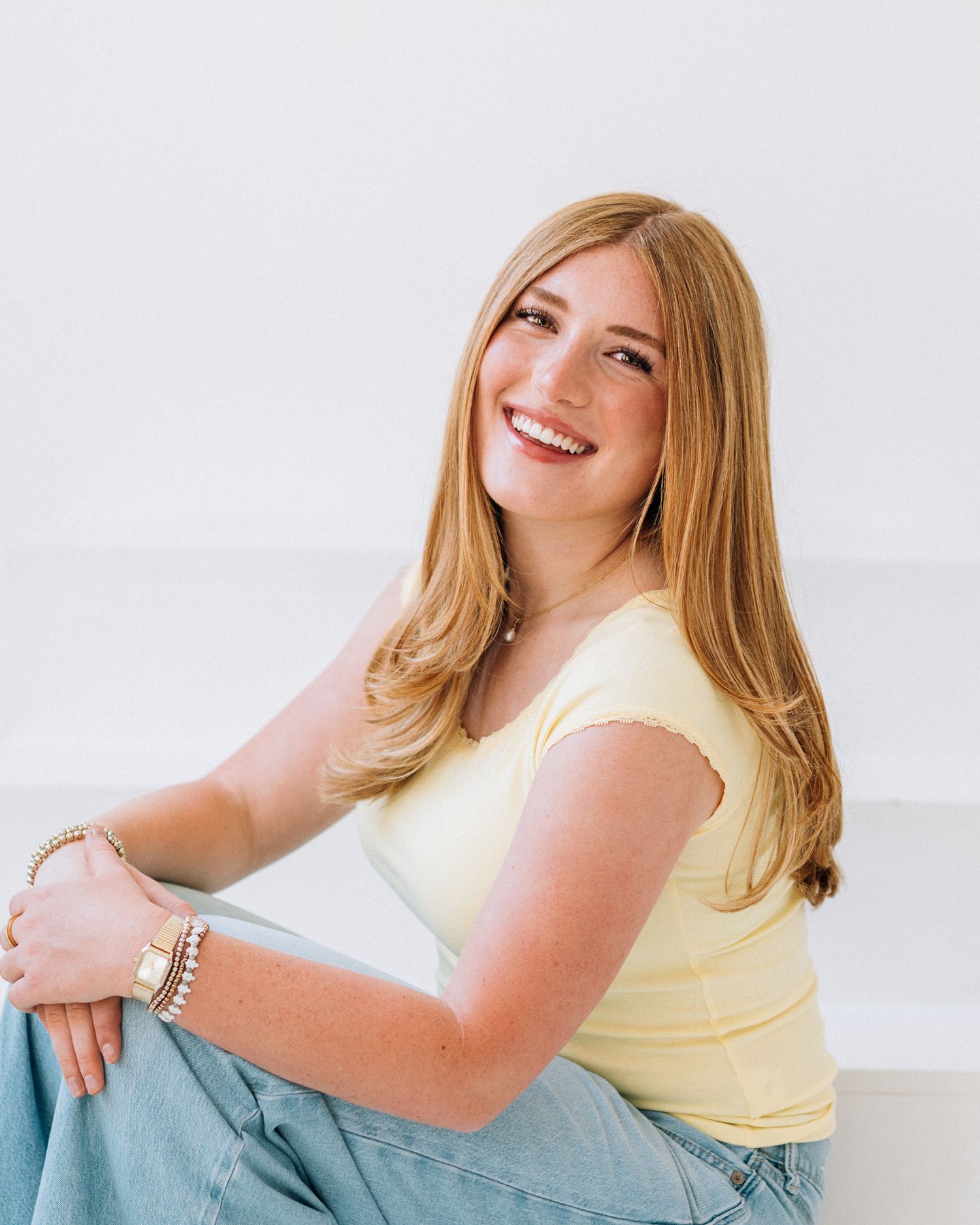 Young woman with long auburn hair smiling, wearing a yellow top and jeans, sitting casually against a white background.