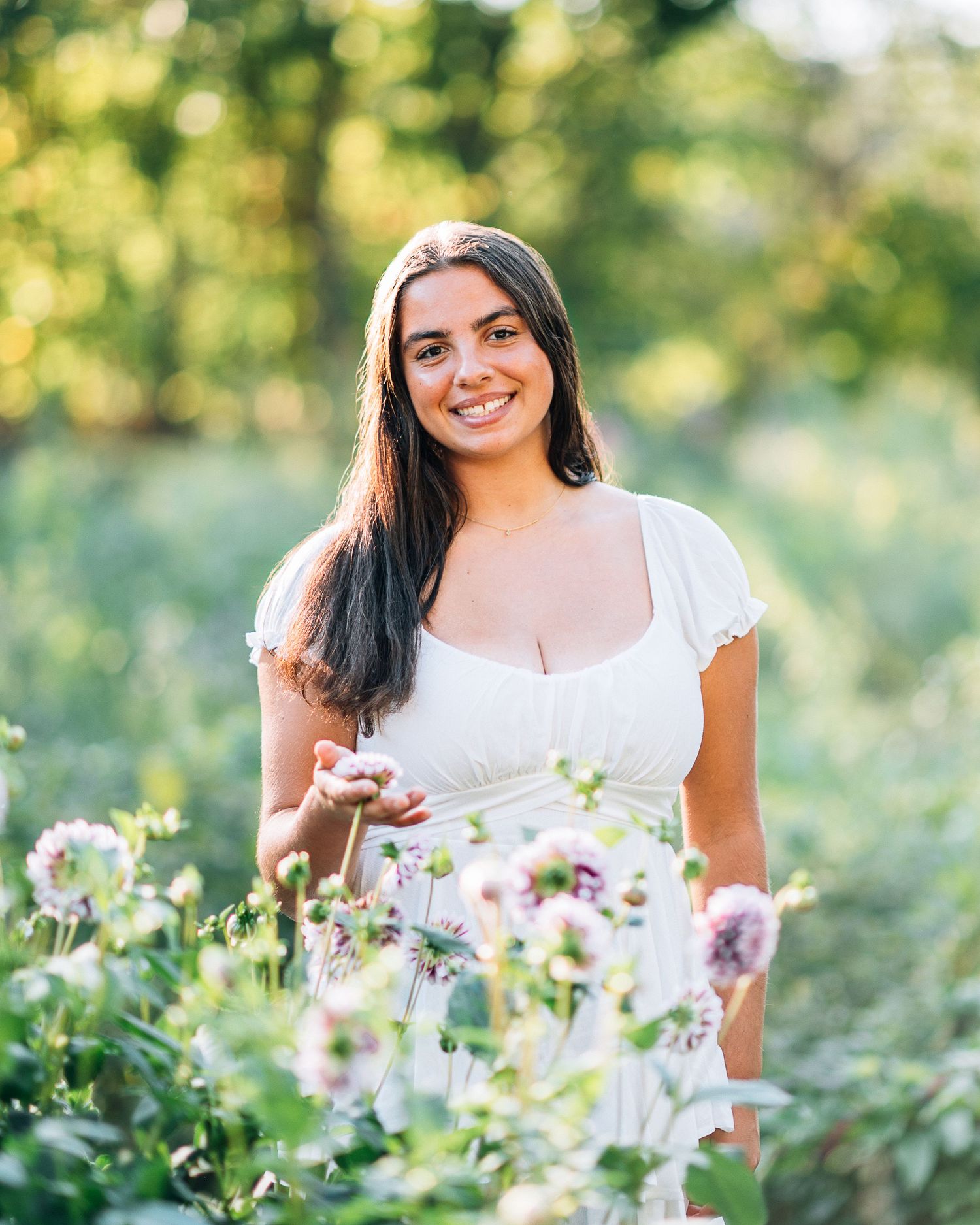 Young woman in white dress smiling among wildflowers in a sunlit garden.