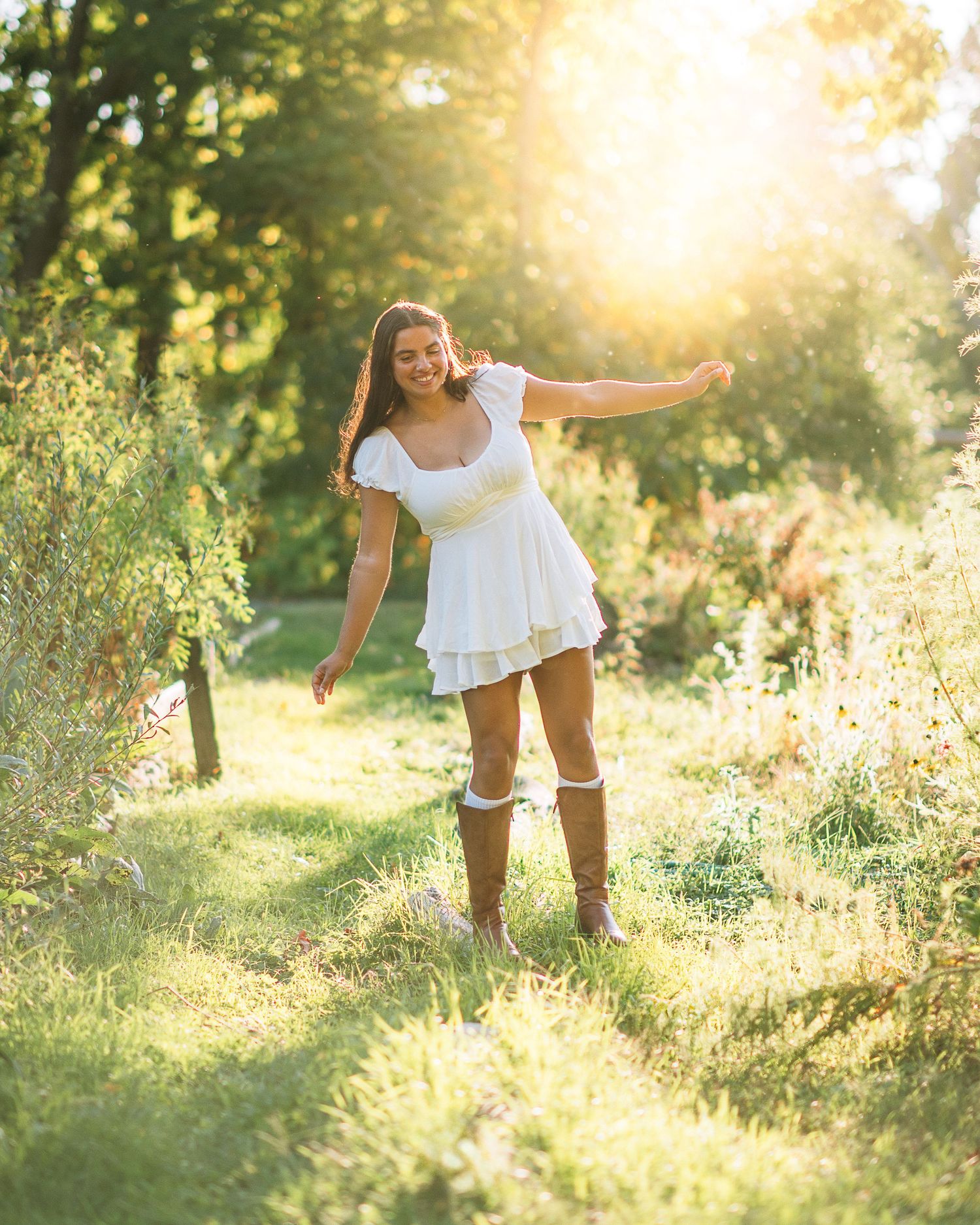 Young woman in white dress and brown boots poses gracefully in sunlit garden with arms outstretched.