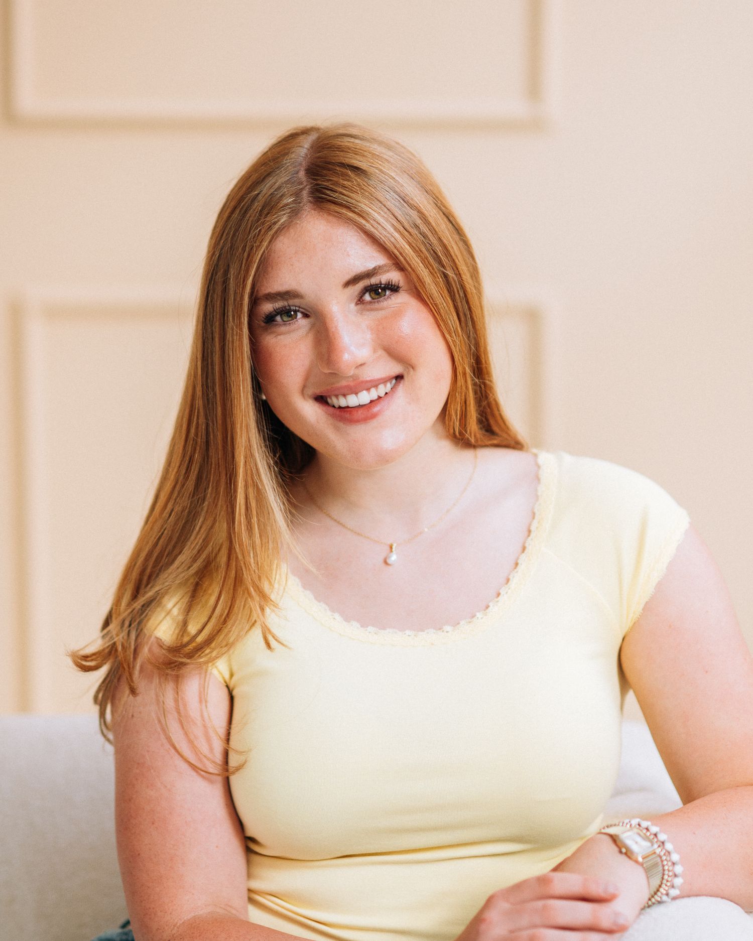 Smiling young woman with long red hair wearing a yellow top, seated in a bright, elegant room.