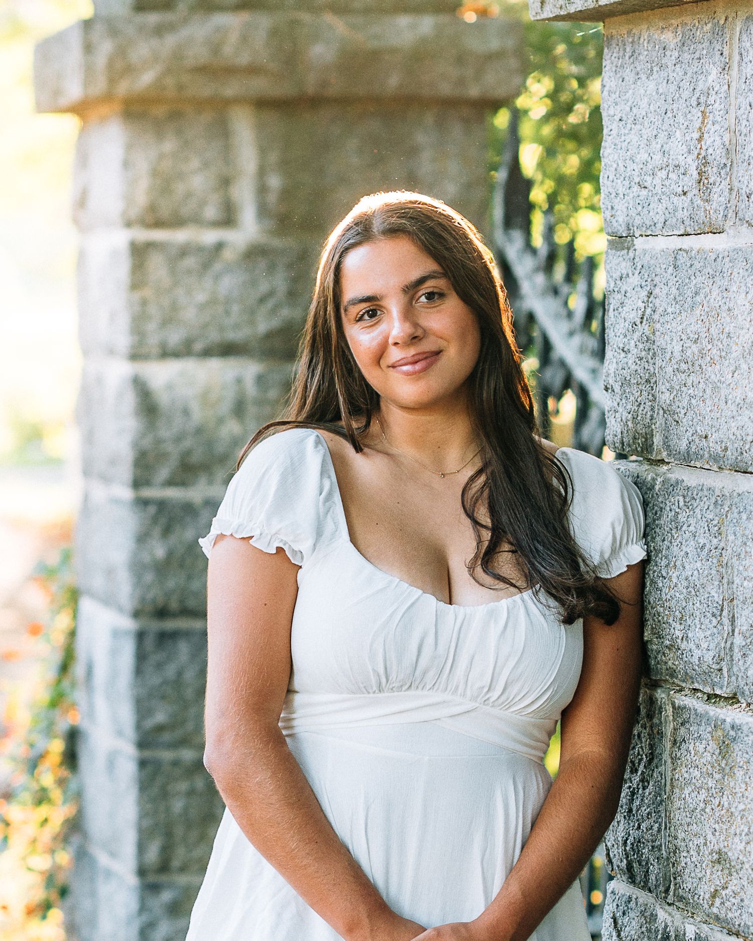 Young woman in white dress smiling against stone wall outdoors in warm sunlight.